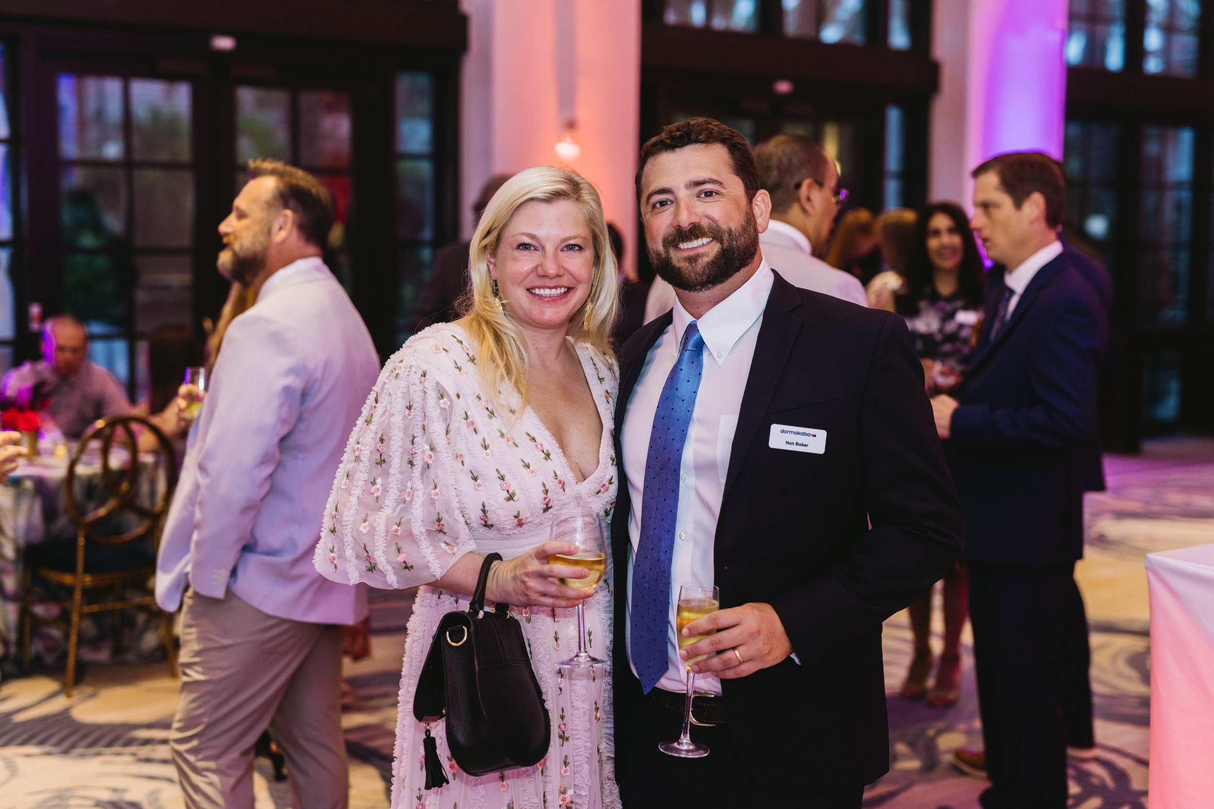 A woman and a man smiling at a social event, holding glasses of champagne. The woman has blonde hair and is wearing a light pink dress with embroidery, carrying a black purse. The man has a beard and is dressed in a dark suit, white shirt, blue tie, 