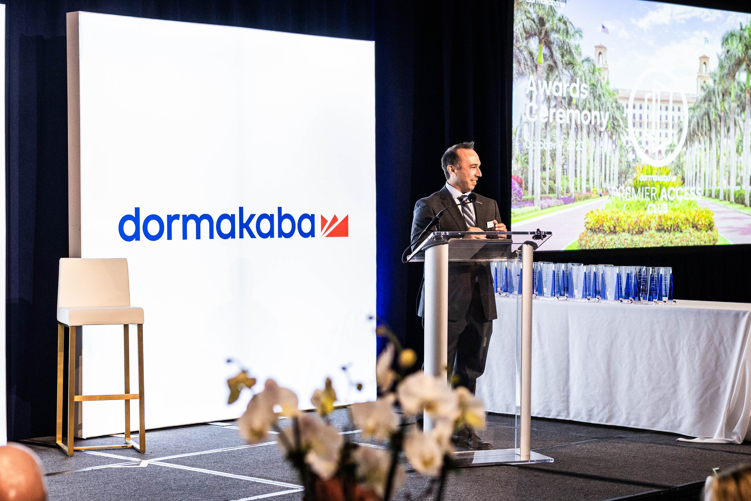 Man in a suit speaking at a podium during an awards ceremony with a large screen displaying a sign that says 'dormakaba' and another screen showing a garden scene with text 'Awards Ceremony October 2023' at The Breakers Palm Beach