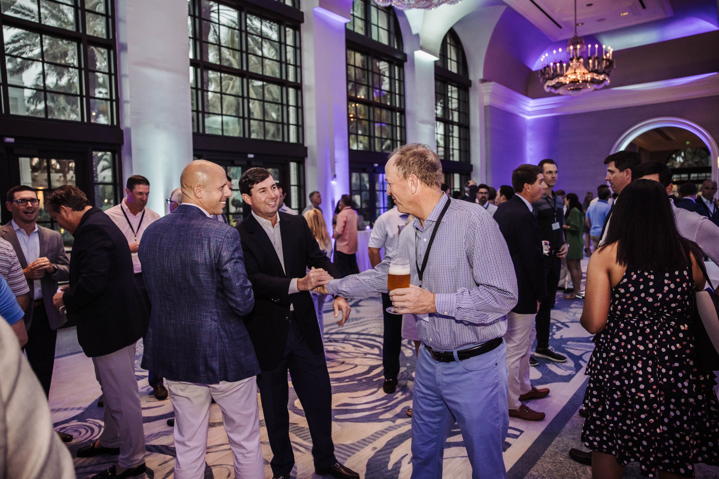 A group of people at a conference or social event in a large, elegant room with tall windows, purple lighting, and chandeliers. Some are talking, shaking hands, and holding drinks. The Breakers Palm Beach, corporate event photography