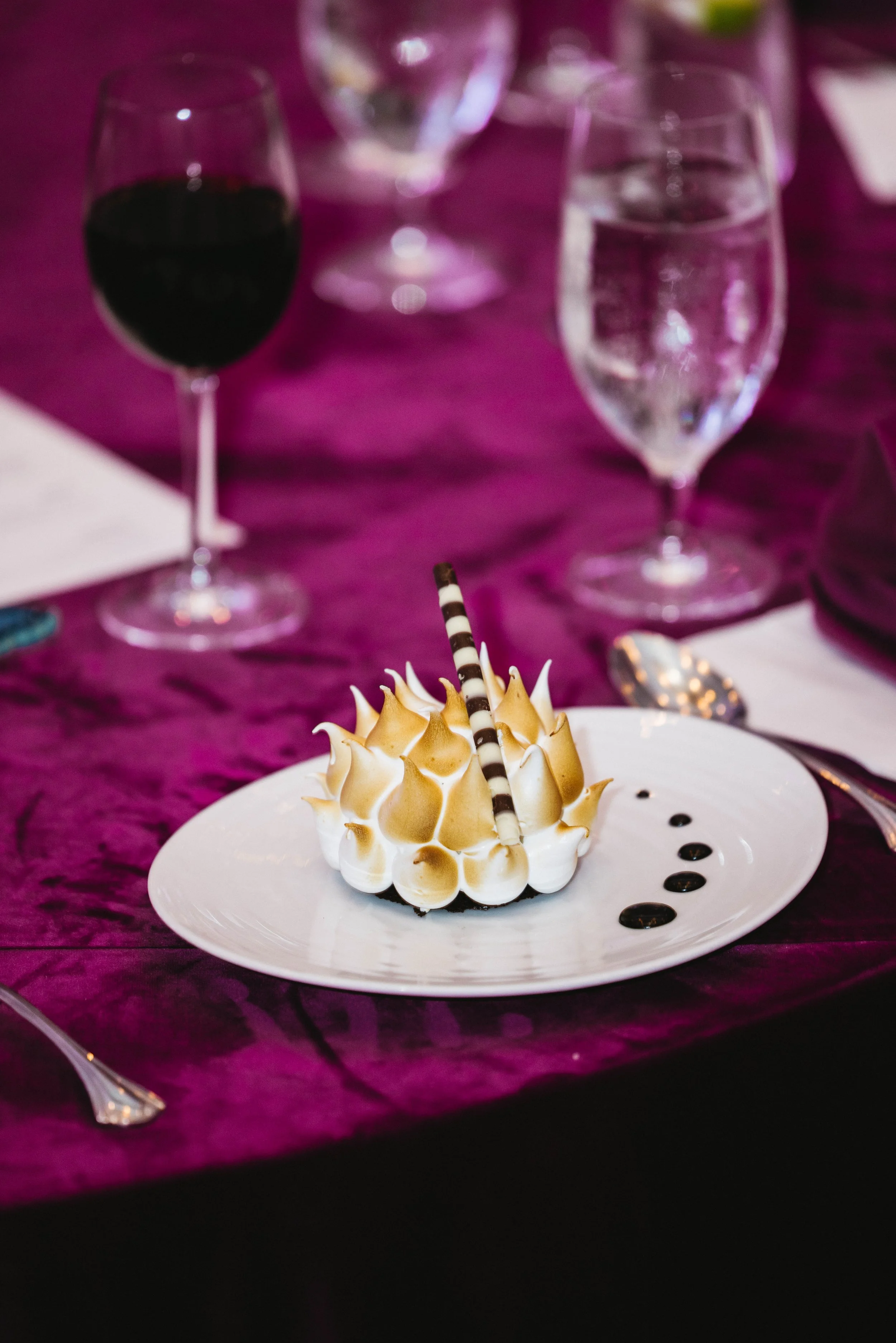 A decorated dessert with whipped cream and a striped chocolate stick on a white plate, surrounded by glasses of wine and water on a purple tablecloth at The Breakers Palm Beach.