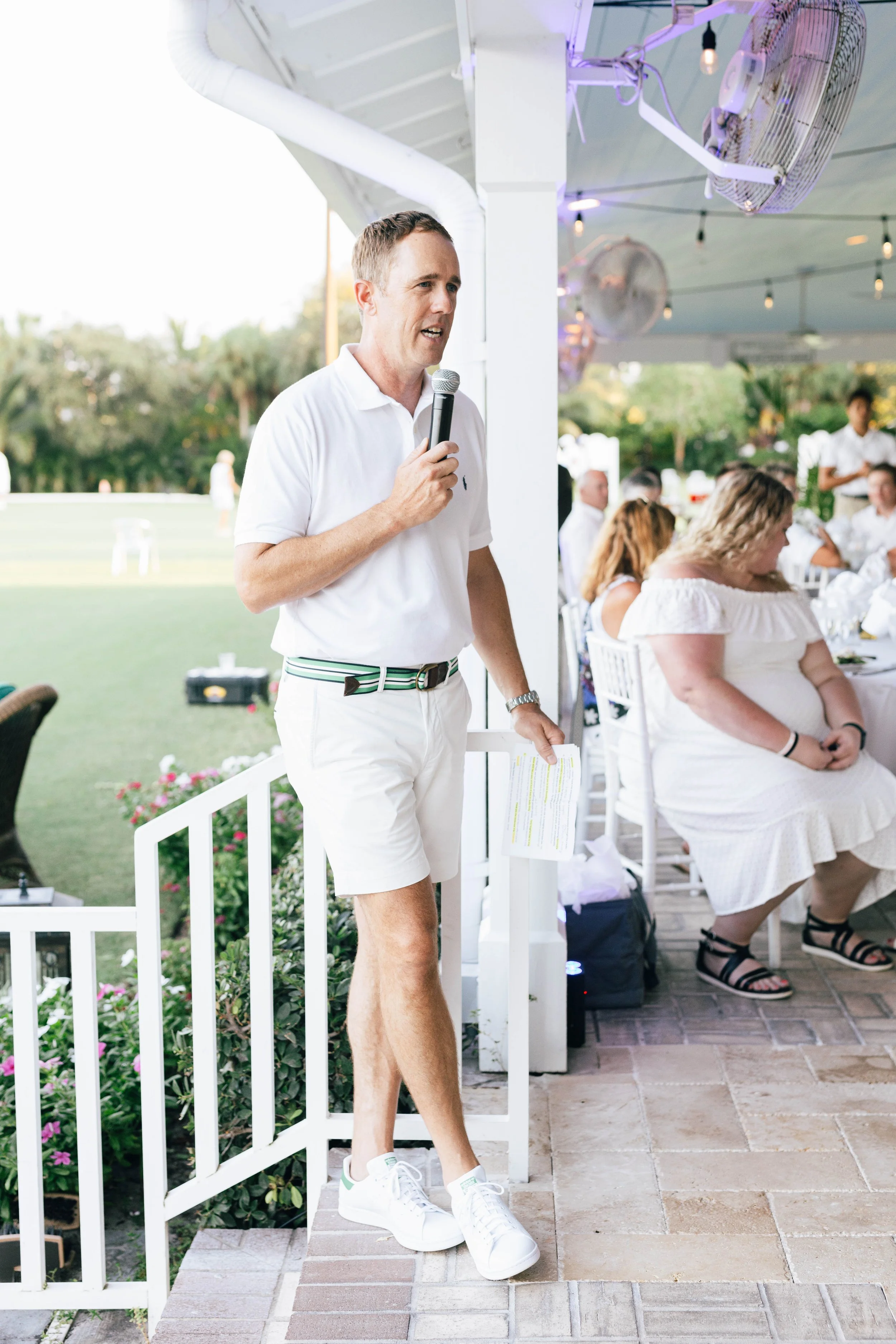 A man in a white polo shirt and shorts giving a speech at an outdoor event, holding a microphone and a paper, with a seated audience in the background. The National Croquet Club, corporate event photography