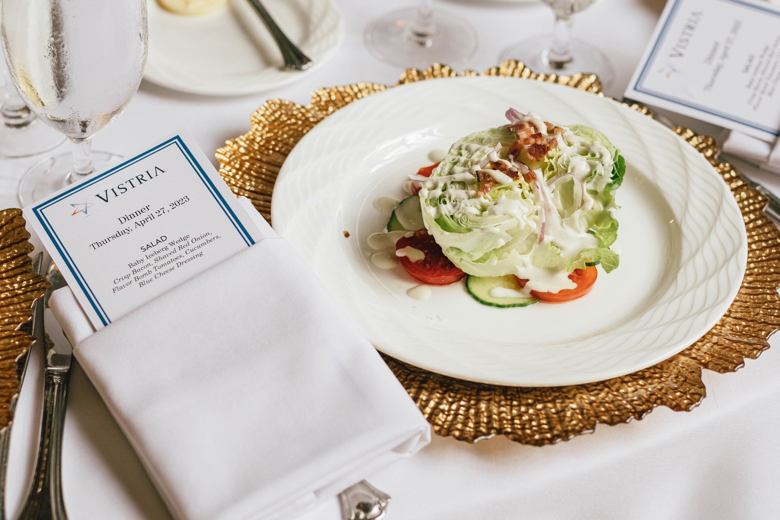 A formal dinner plate with a fresh green salad topped with dressing, placed on a gold charger plate. To the left, there is a folded white napkin with a printed dinner menu. The setting is on a white tablecloth.  The Breakers corporate event photo