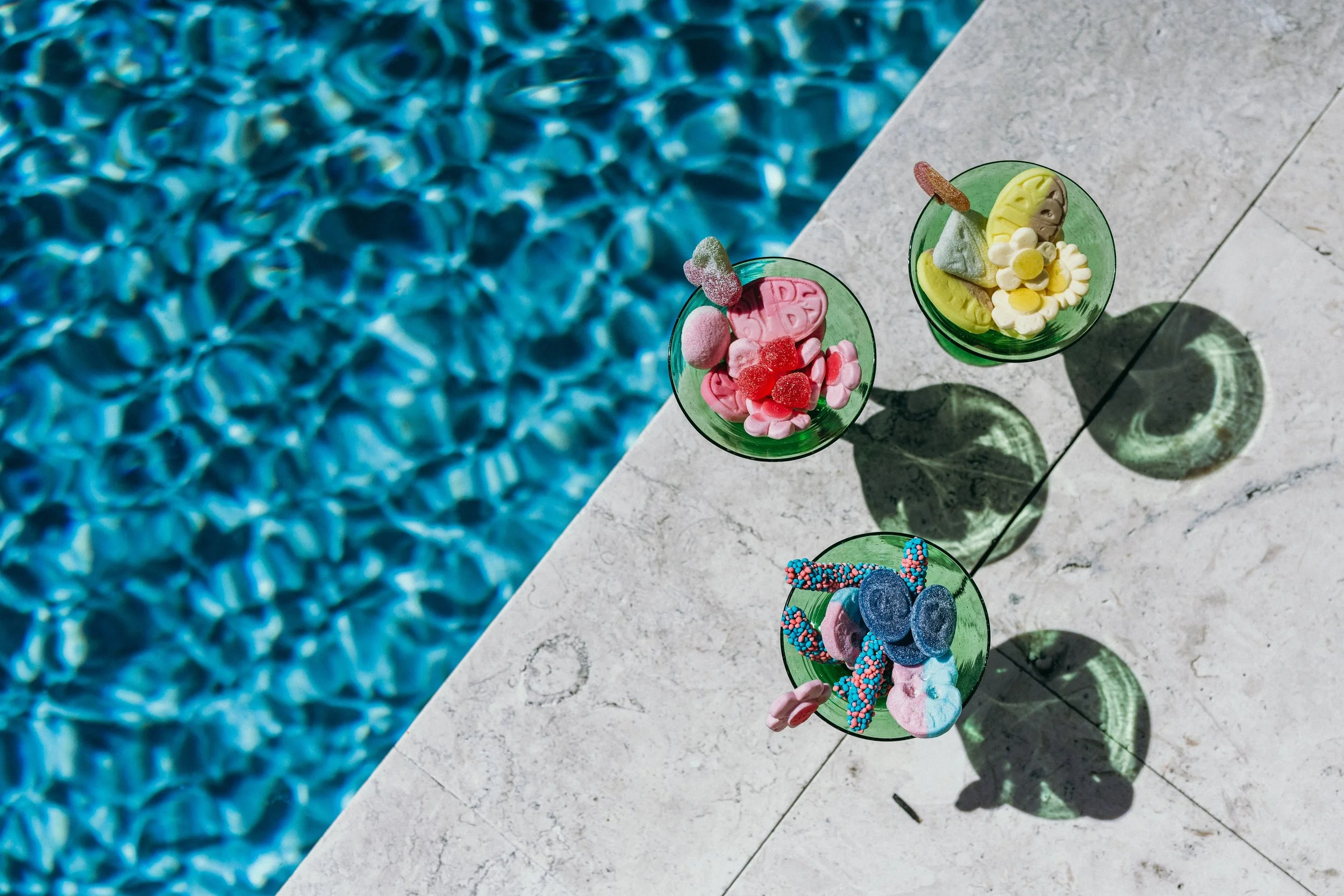 Three green glasses containing colorful candies placed on a light gray stone surface beside a pool. Brand activation photography.