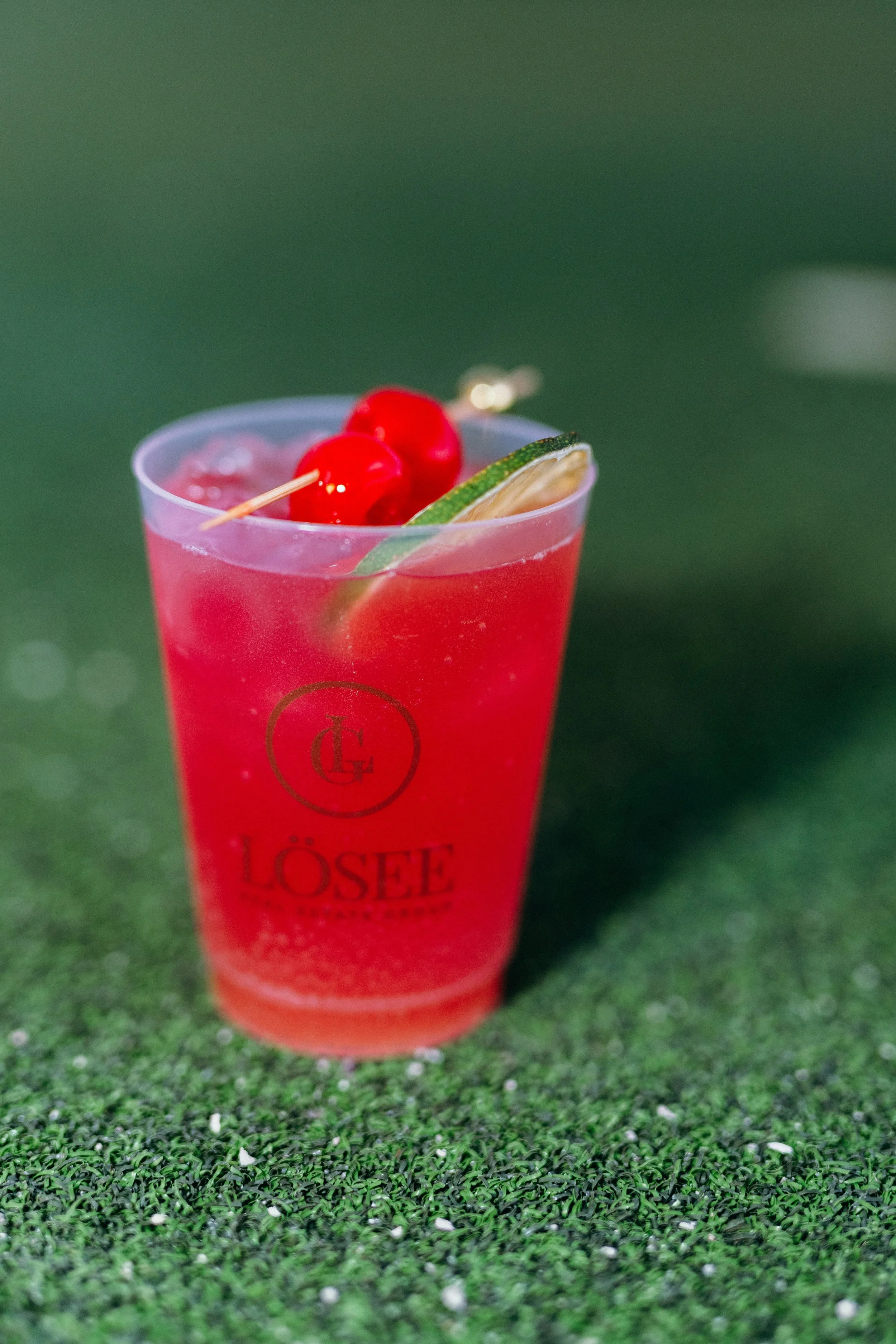 A red cocktail garnished with cherries and a lime wedge in a clear plastic cup on a green surface.