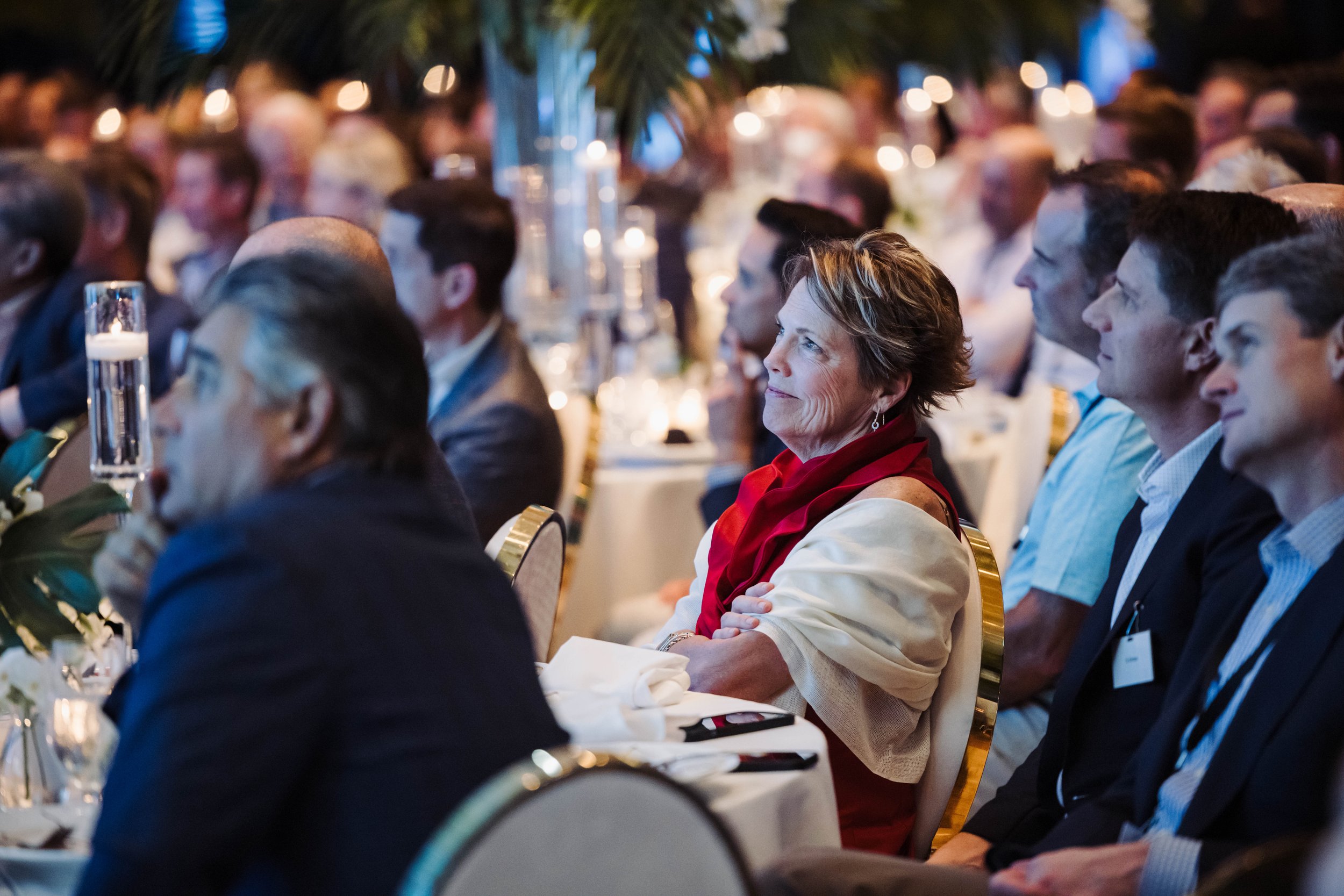 Attentive audience at The Breakers Palm Beach seated at round tables during a formal event, with candles and floral arrangements, with a woman in a red scarf in focus.
