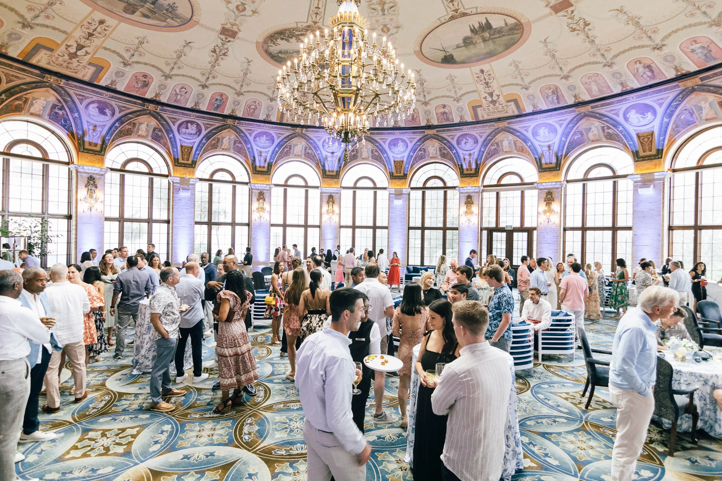 A large, elegant hall filled with people socializing, dining, and mingling during a corporate event at The Breakers Palm Beach. The hall has tall, arched windows, decorative painted ceilings, and a grand chandelier hanging from the center.