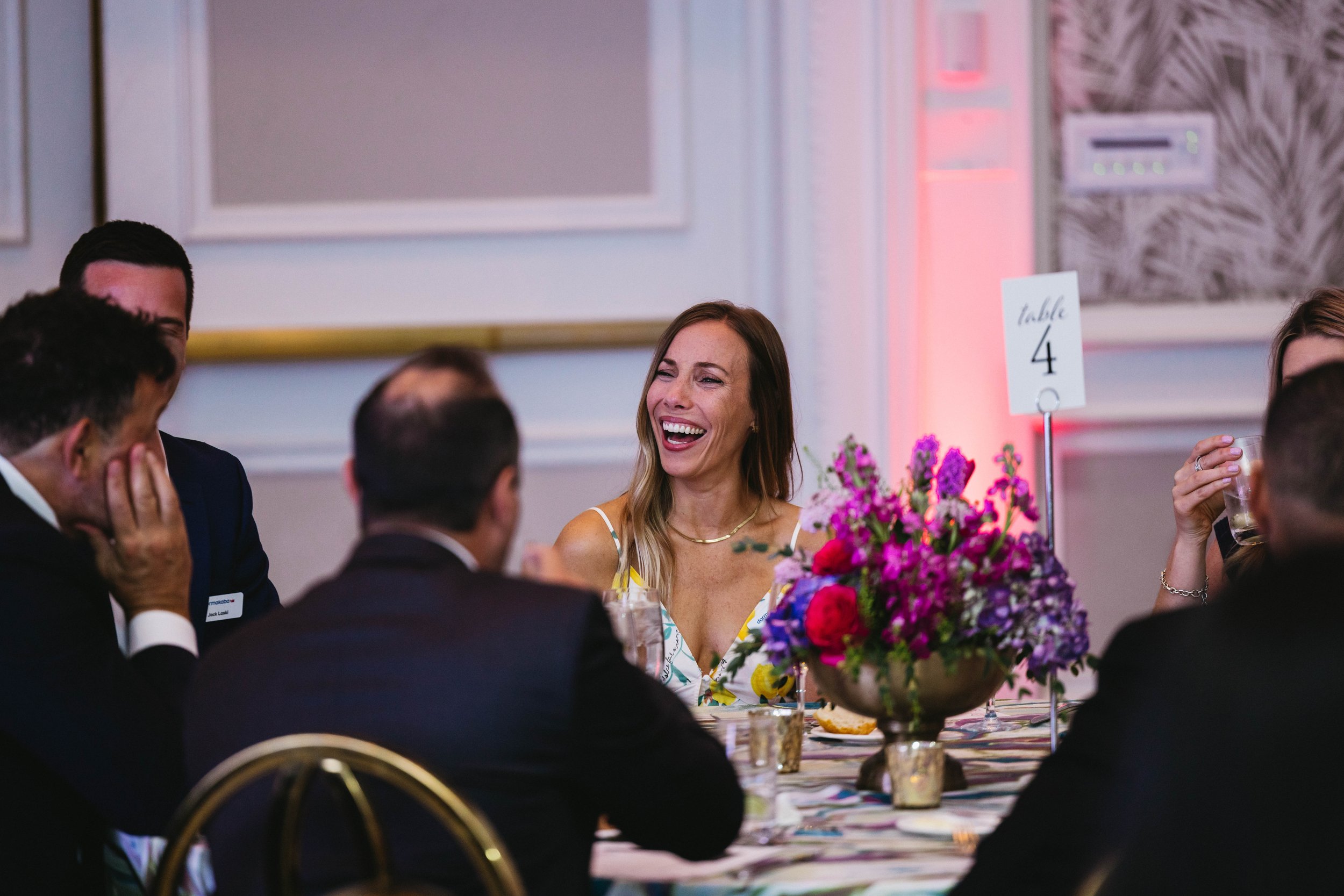 A woman with long blonde hair, wearing a sleeveless dress, is laughing at a formal dinner event surrounded by men in suits. They are seated at a table with a colorful floral centerpiece, glasses, and table number sign at The Breakers Palm Beach.