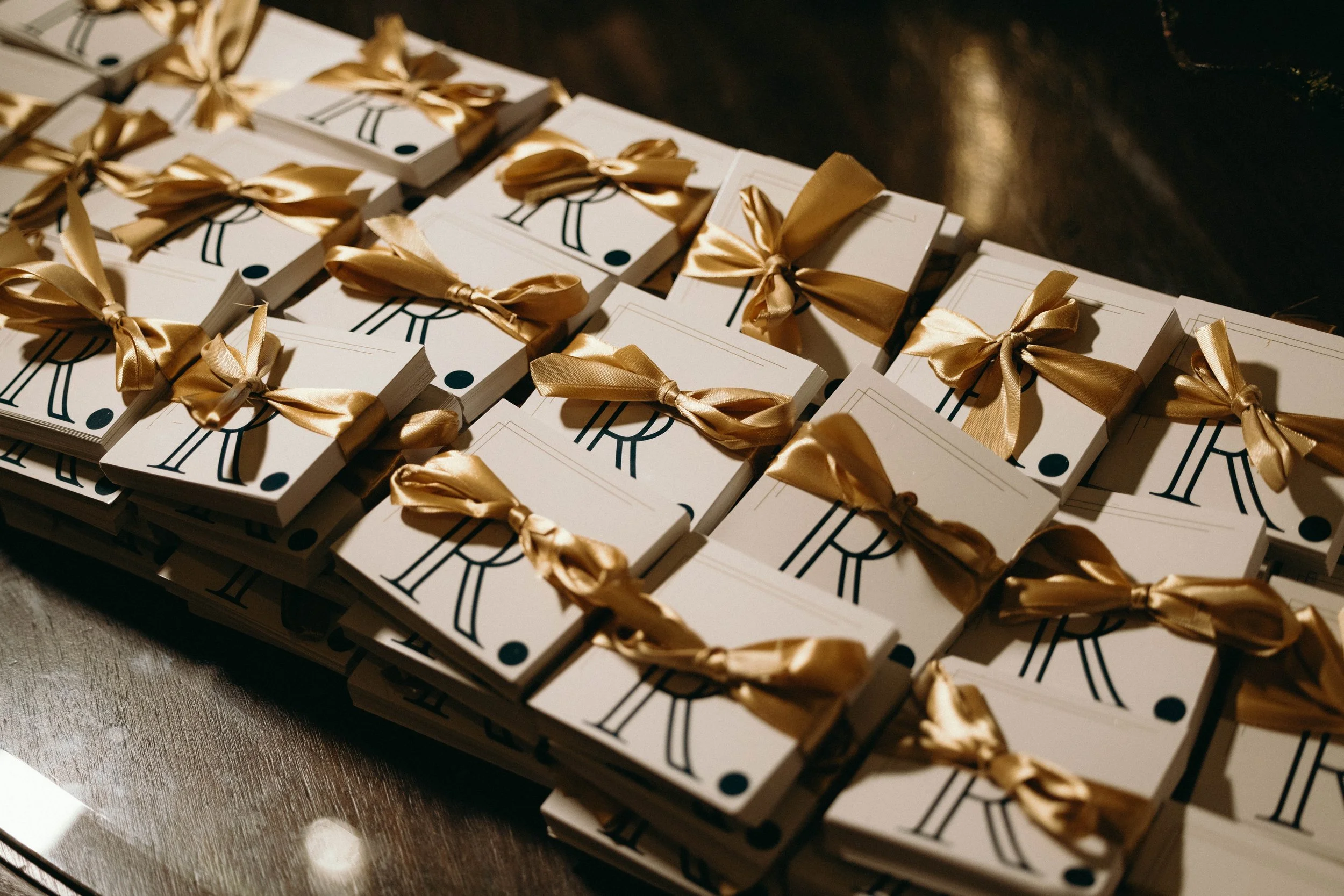 Multiple small white boxes with a stylized clock face and a black dot, tied with gold satin ribbons, arranged on a dark wooden surface at The Boca Raton hotel.
