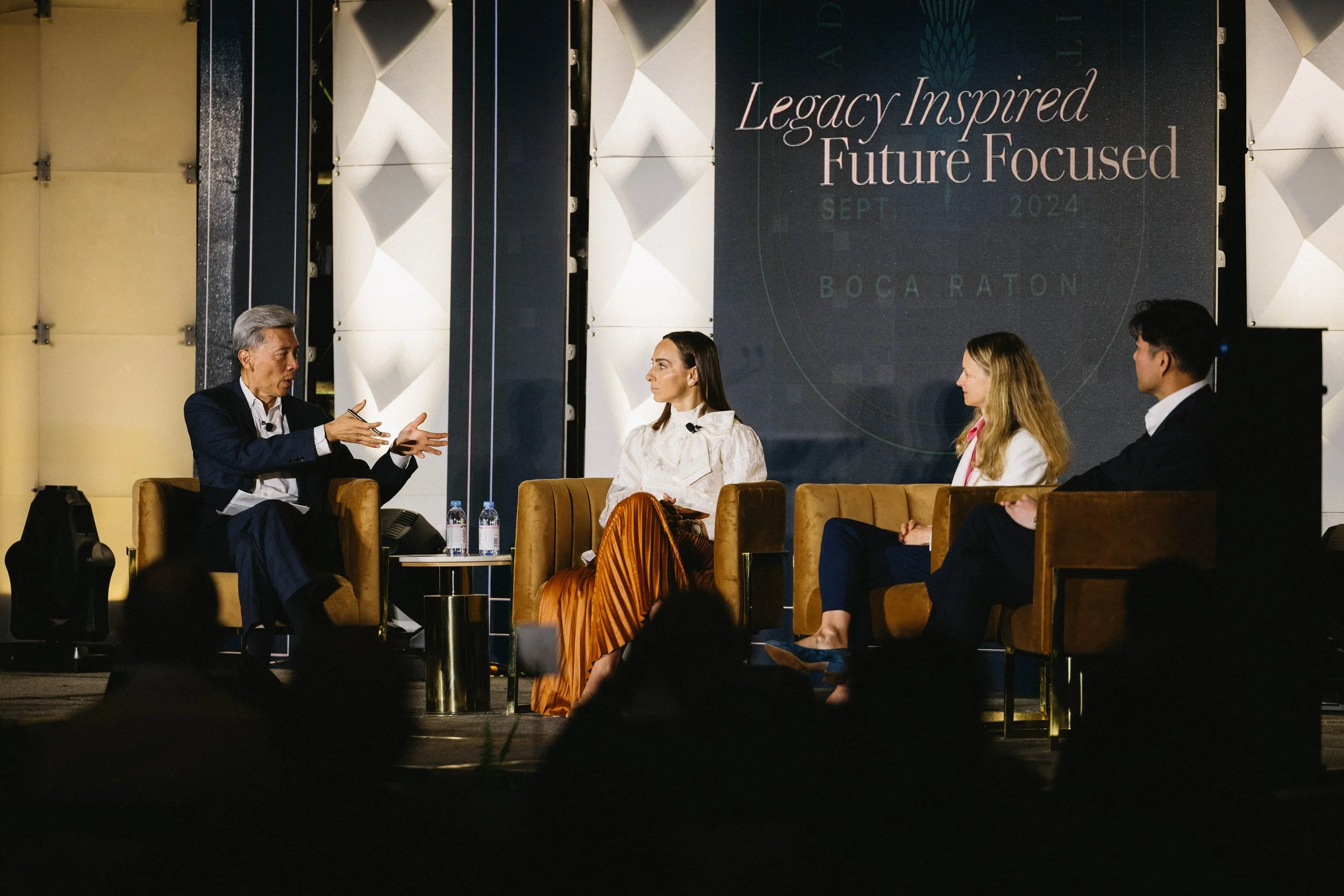 Four people sitting on stage in a panel discussion at an event titled 'Legacy Inspired Future Focused' with a large screen behind them displaying the event details. The Boca Raton Hotel, corporate event photography