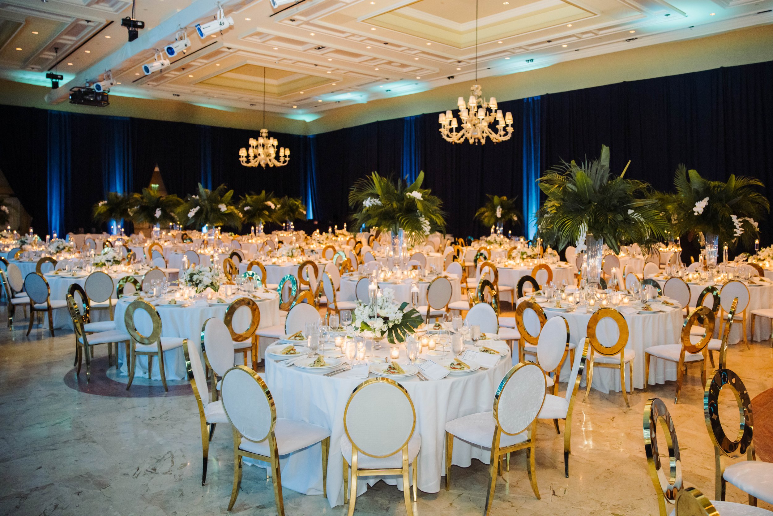 Elegant banquet hall decorated with round tables covered in white tablecloths, gold and white chairs, tall floral centerpieces with greenery, and candles, with dark curtains and chandeliers overhead at The Breakers Palm Beach.