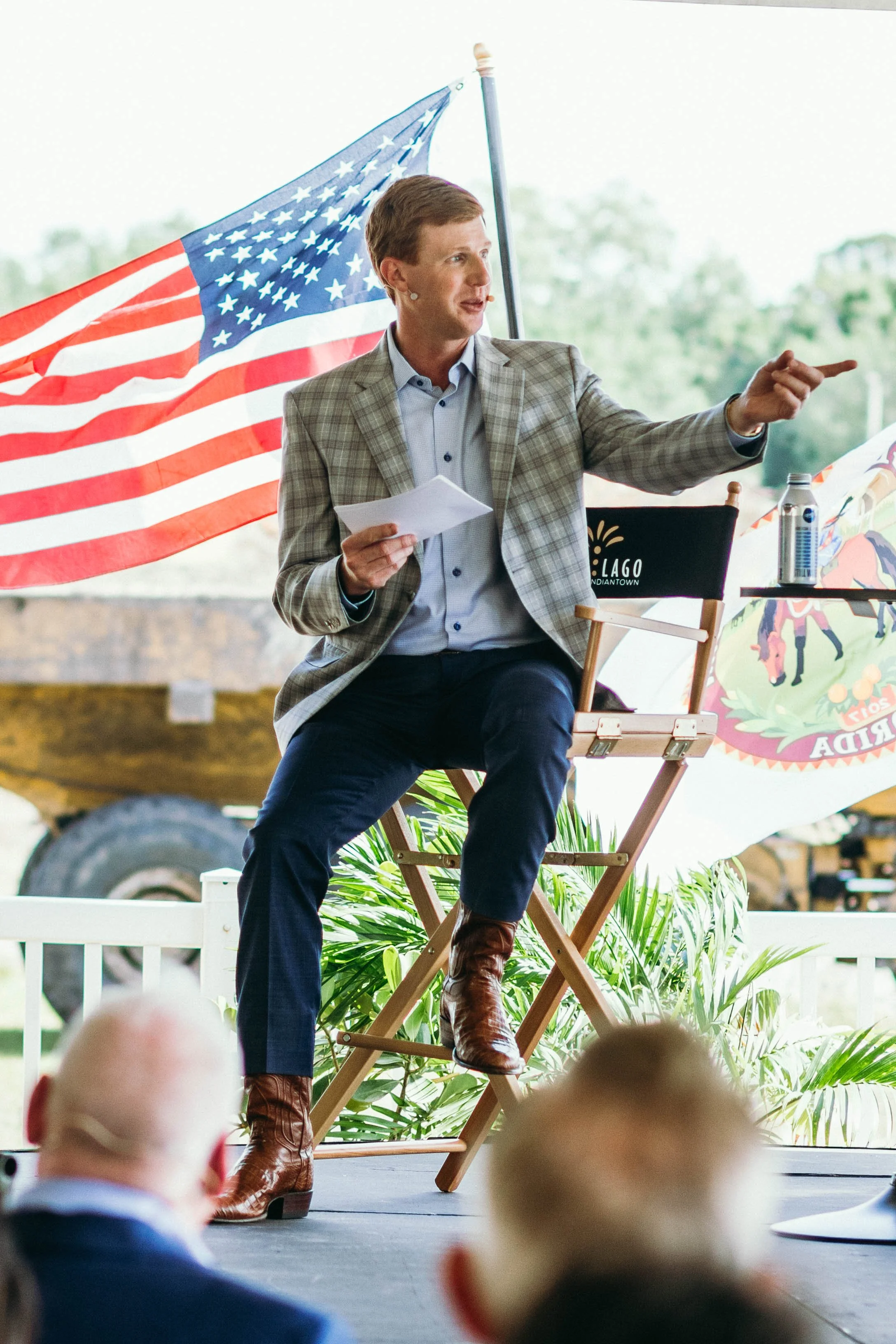 A man sitting on a director's chair, wearing a checked blazer, blue shirt, and brown boots, gesturing with his right hand while holding papers in his left hand at an outdoor event with American and Florida state flags in the background.