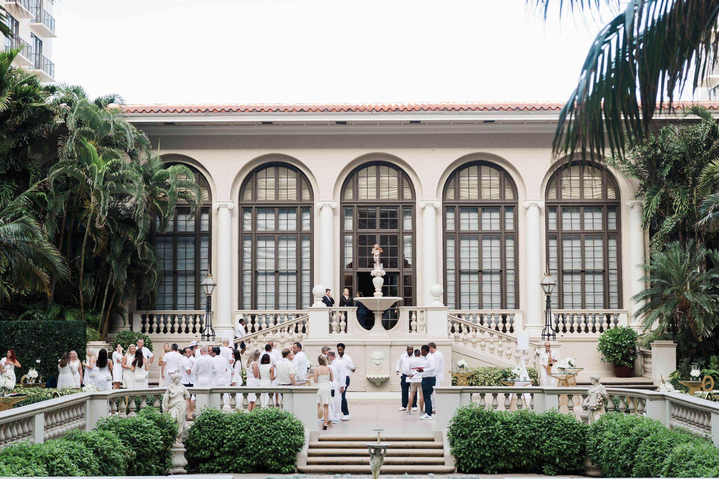 A corporate event at The Breakers Palm Beach with large arched windows, a fountain, and lush greenery, with people dressed in white and formal attire.