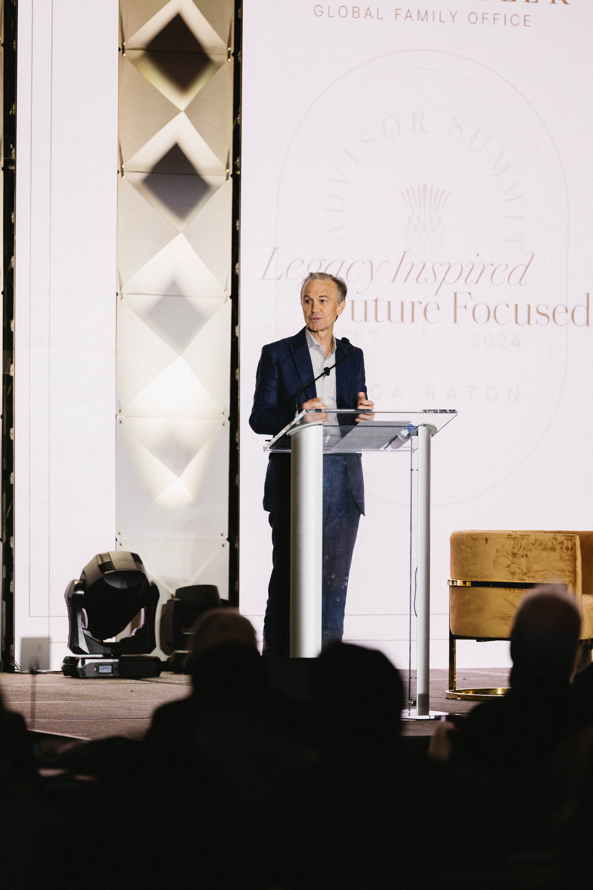 A man in a dark suit stands at a podium speaking at a conference, with a large screen behind him displaying the event's theme and logo. The Boca Raton Hotel, corporate event photography