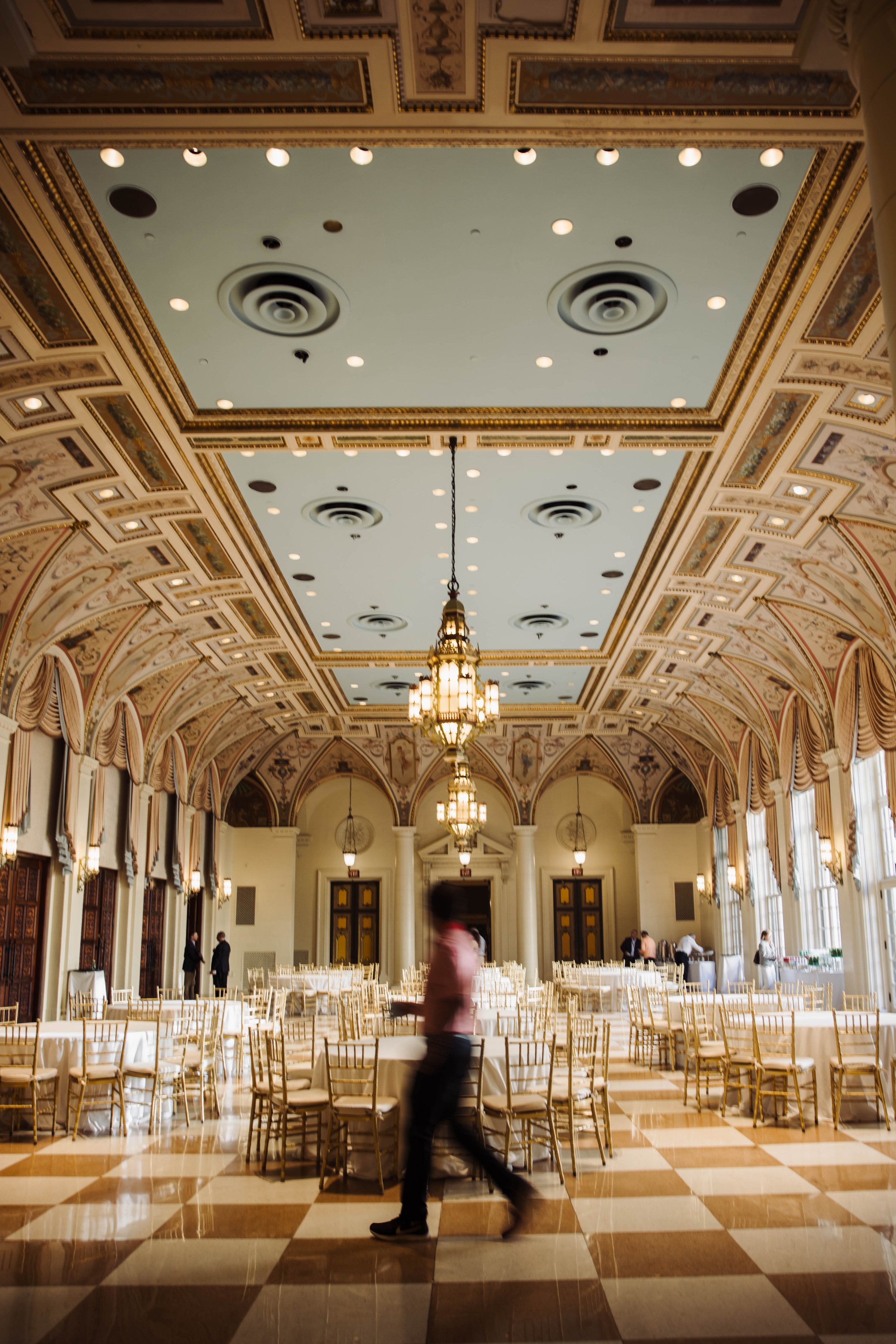 Elegant banquet hall with ornate ceiling, chandeliers, large windows, round tables with white tablecloths, gold chairs, and a few staff members preparing the room at The Breakers Palm Beach. Corporate event photography.