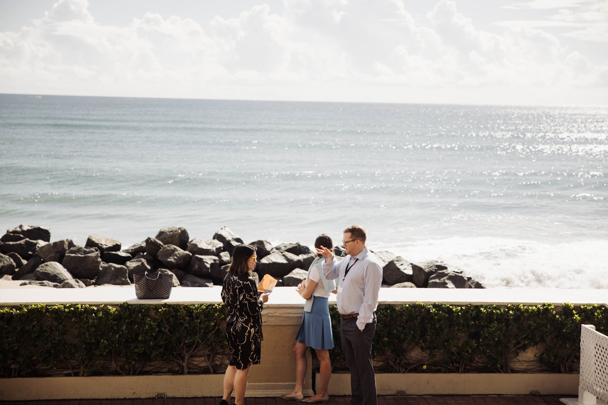 Three people stand by a seaside railing with rocks and ocean in the background, one woman reads a book, another woman stands with her back to the camera, and a man in glasses gestures with his hand at The Breakers Palm Beach.