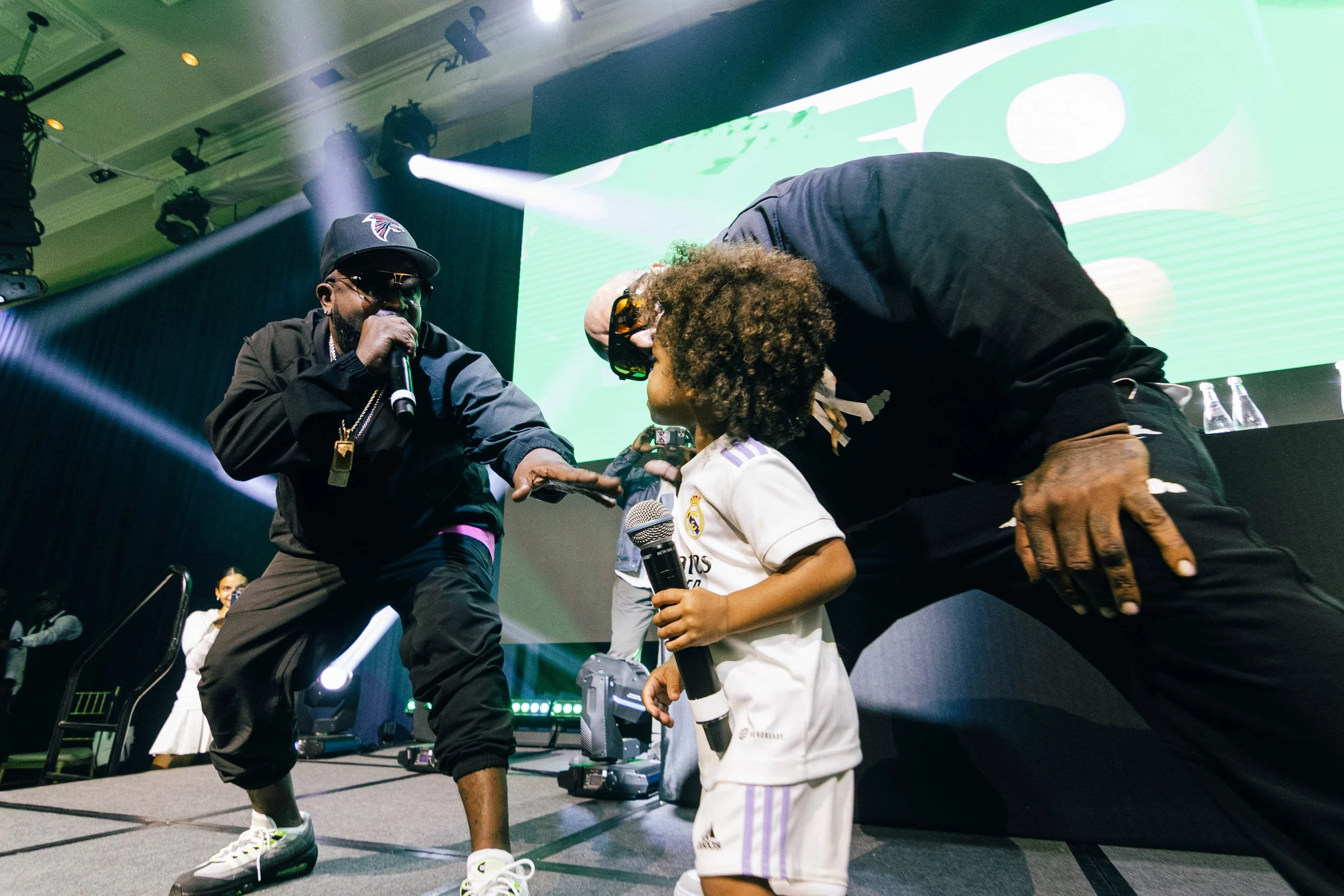 Big Boi from Outkast and Sleepy Brown on stage during a concert or event, with the performer engaging with the child who is holding a microphone, against a backdrop with colorful lights and large digital screen. Corporate event photography.