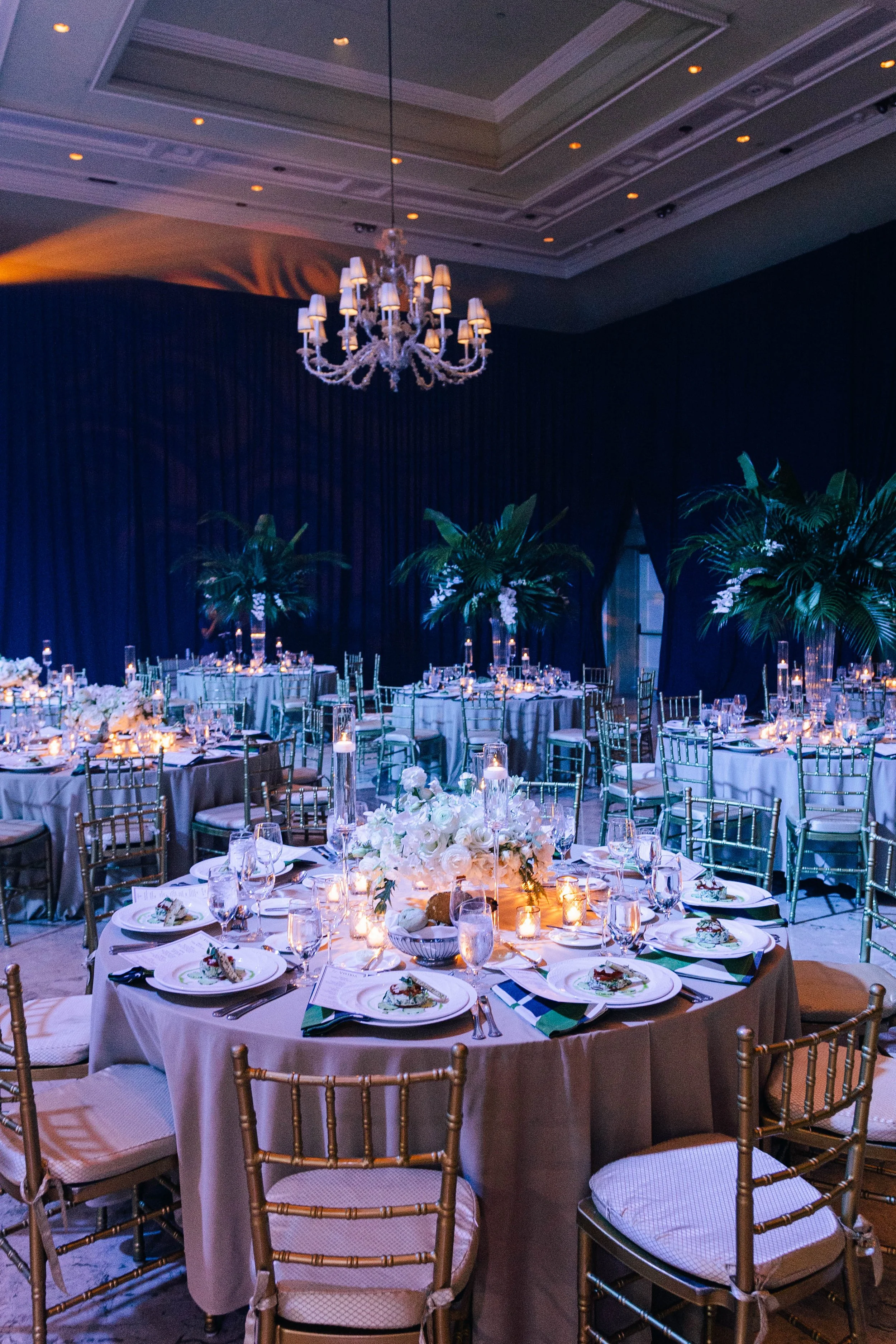 Elegant banquet hall at The Breakers Palm Beach decorated with round tables covered in beige tablecloths, set with glassware, plates, and candles, with lush green plants as centerpieces and a chandelier hanging from the ceiling.