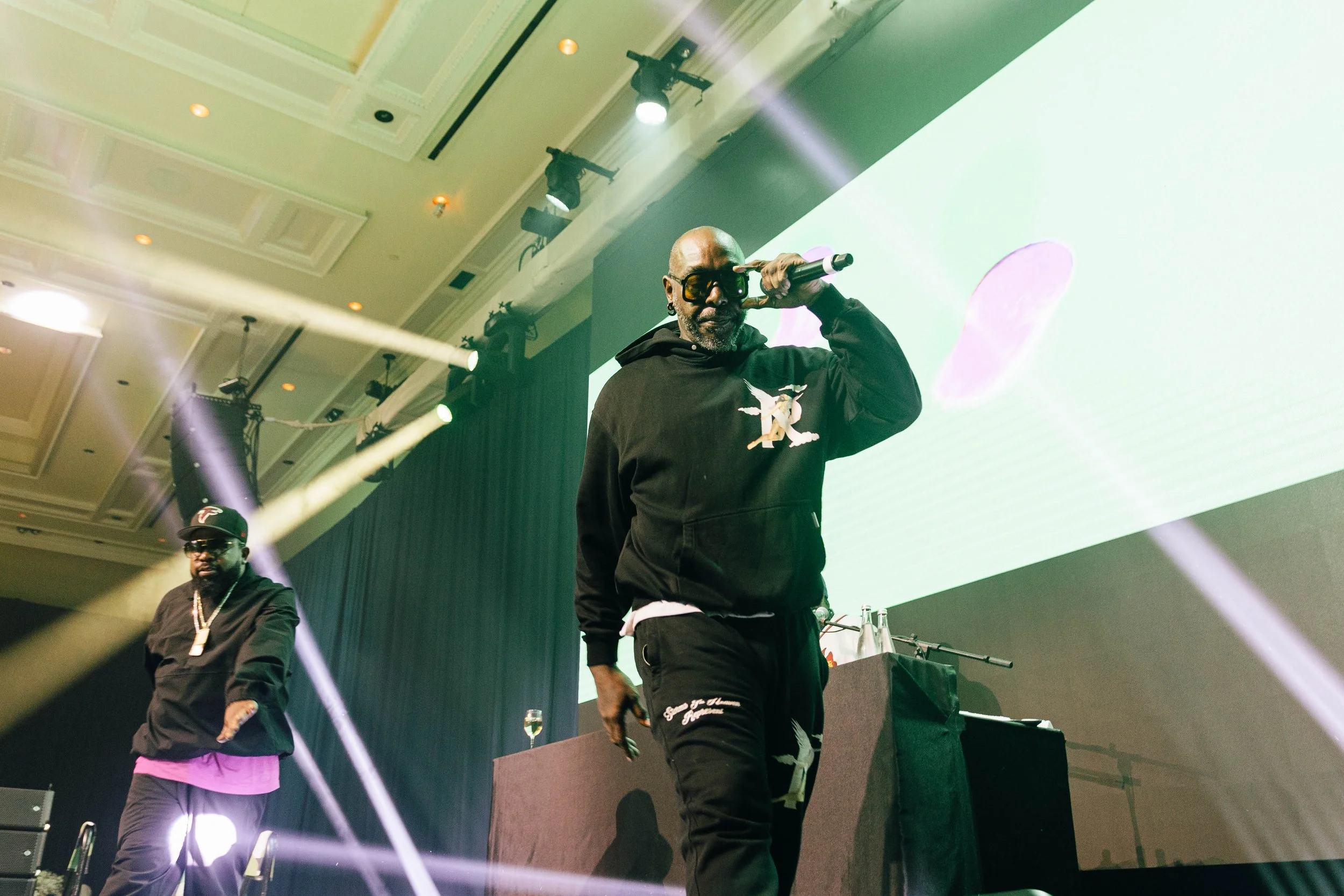 Sleepy Brown, adjusting glasses with one hand and holding a microphone, wearing a black hoodie with a logo, and another in the background in dark clothing with a cap. The Breakers Palm Beach,  corporate event photography