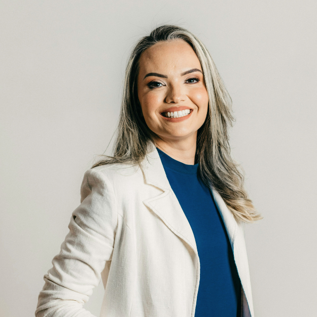 A smiling woman with long blonde hair in a white blazer and blue top standing against a plain white background.