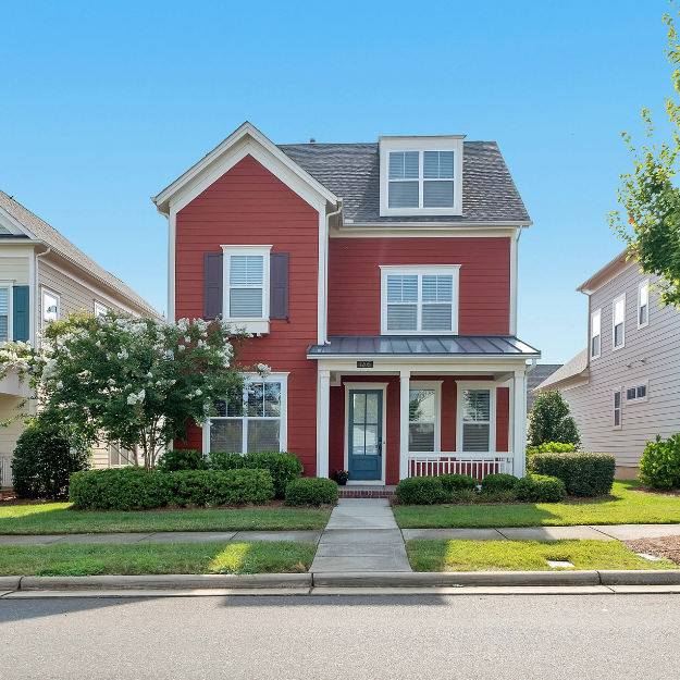 Red three-story house with front porch, surrounded by a well-maintained lawn and shrubs, under a clear blue sky.