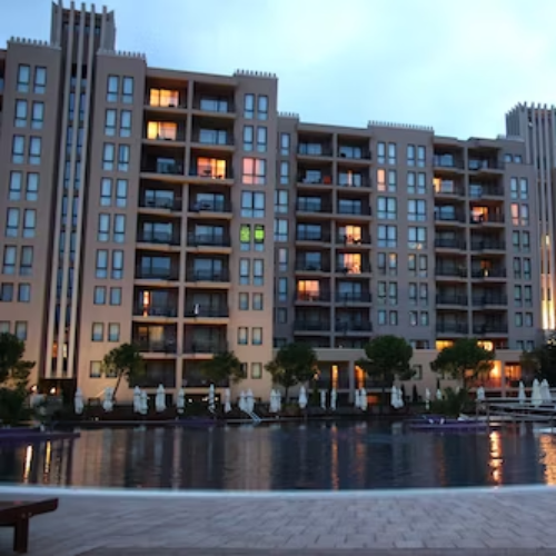 A multi-story apartment building with balconies, illuminated windows, and a waterfront view with trees and patio umbrellas in the foreground, taken during dusk.