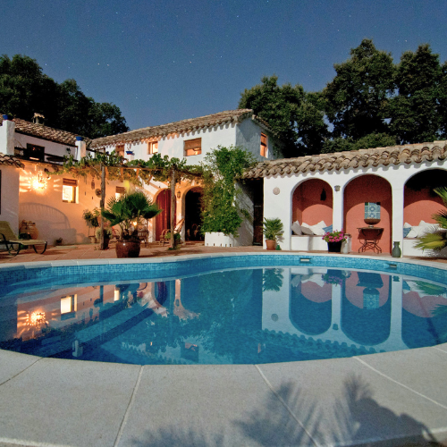 A nighttime scene of a backyard with a swimming pool, Mediterranean-style white and pink villas, palm trees, and outdoor seating under string lights.