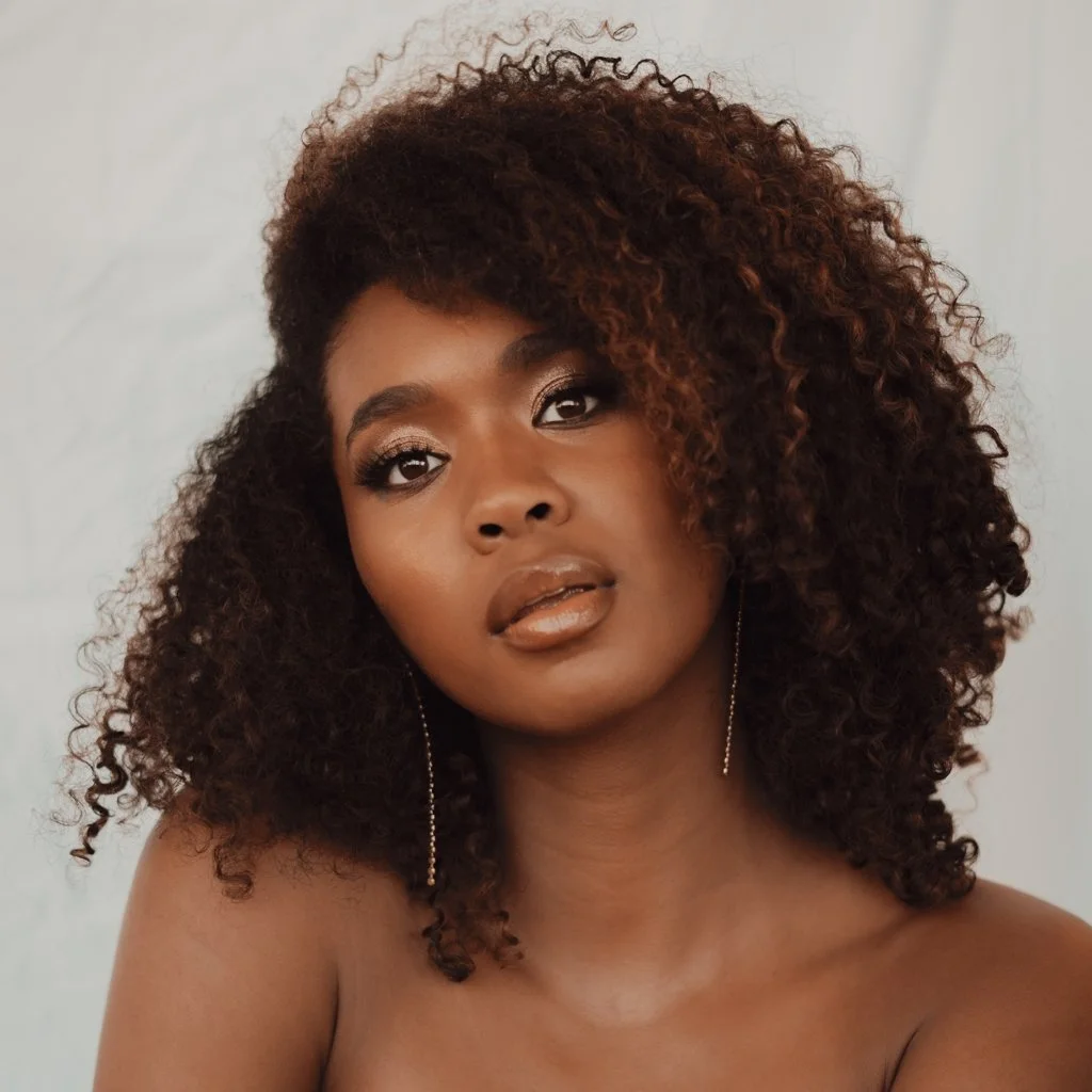 Portrait of a woman with dark skin and curly, voluminous hair. She has makeup with shimmer eyeshadow, eyeliner, and nude lipstick, and is wearing long, dangling earrings.