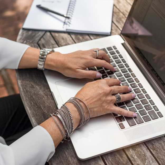 Person typing on a laptop with a notebook and pen on a wooden table.