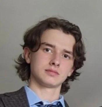 Close-up of a young man with medium-length wavy brown hair, wearing a gray sweater over a light blue shirt and a dark blue bow tie, against a neutral background.
