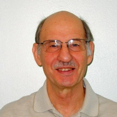 Older man with glasses and a mustache smiling, wearing a light-colored polo shirt, against a plain white wall.