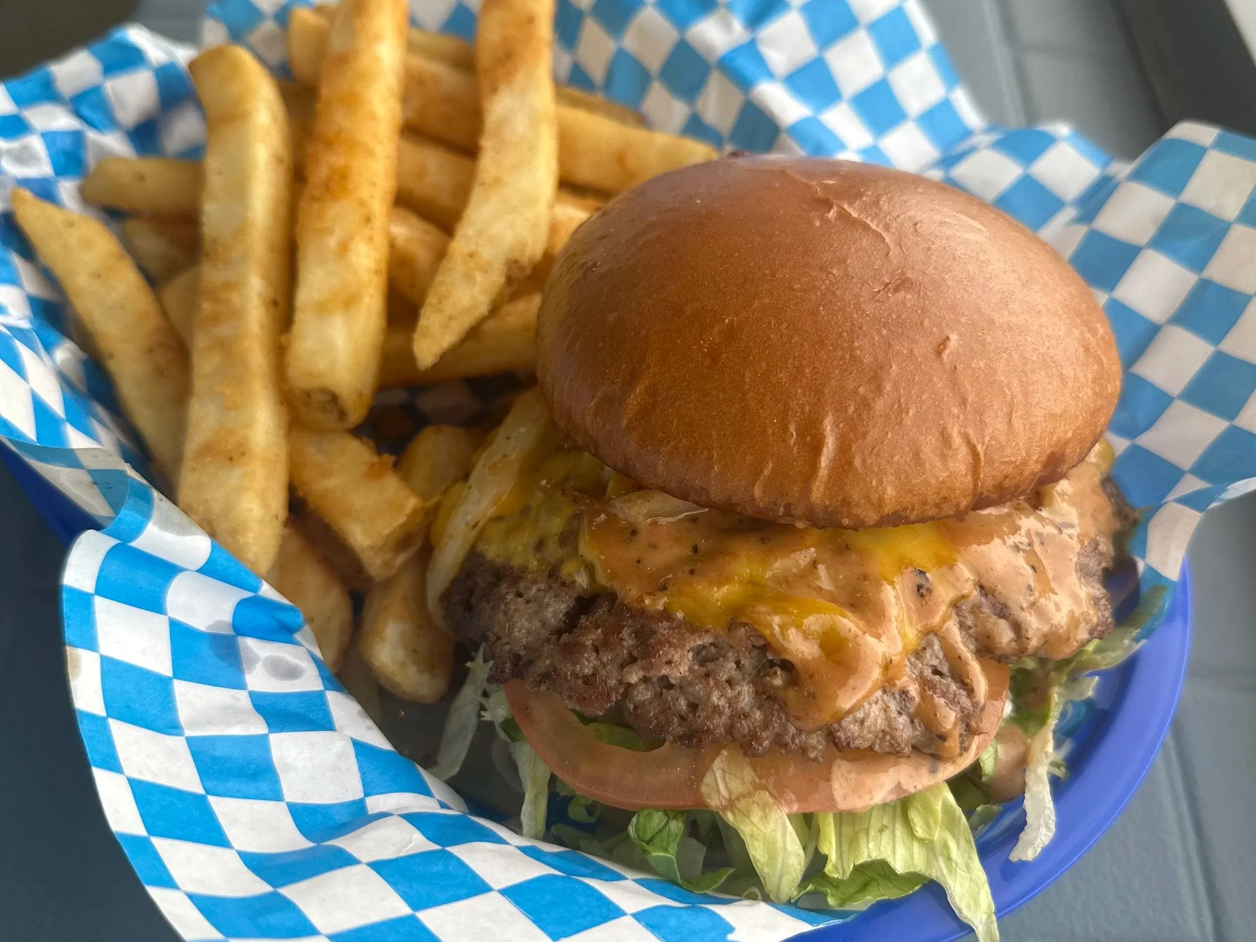 Burger with lettuce, tomato, cheese, and bun served with French fries in a checkered paper-lined basket.