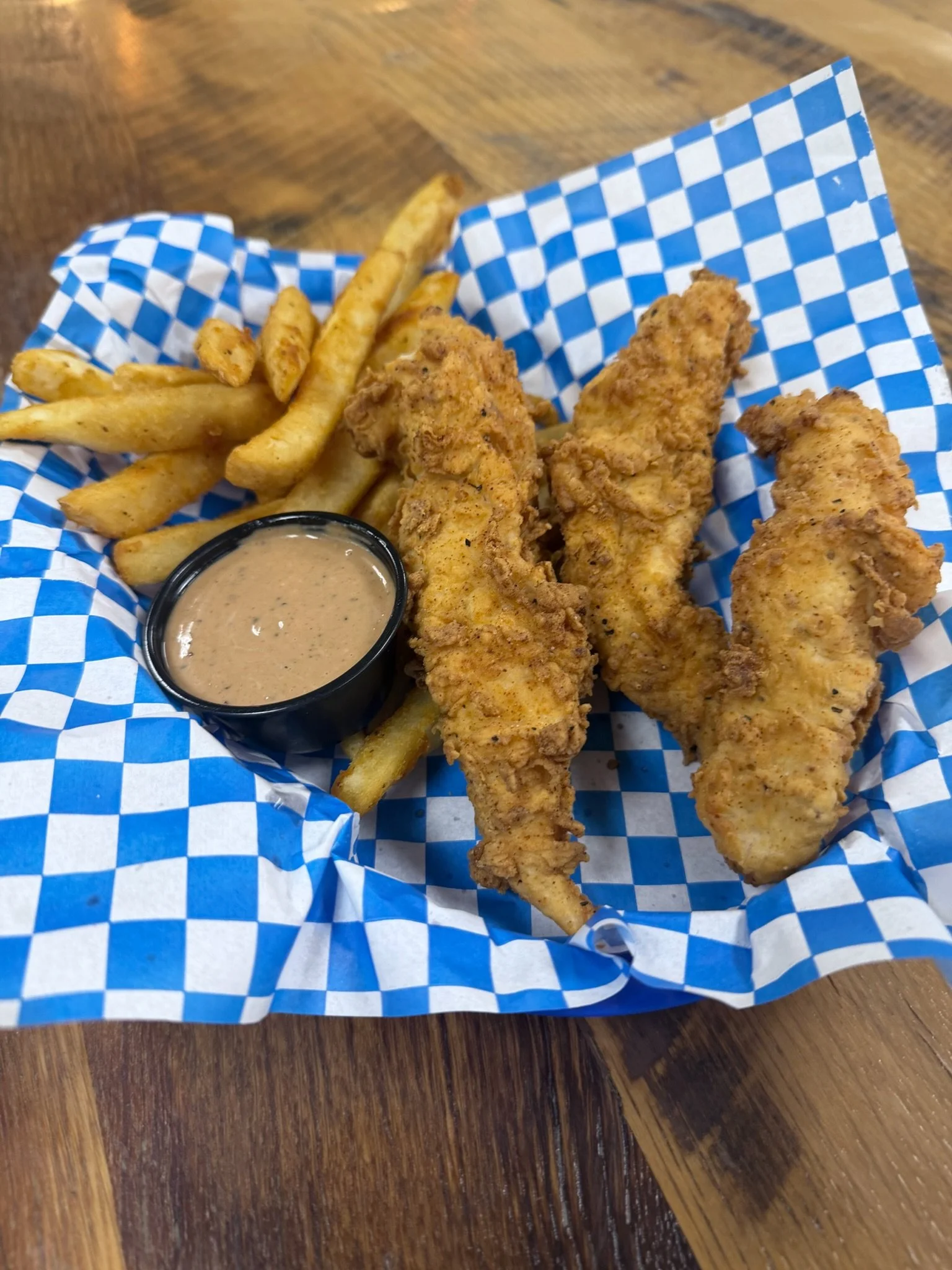 Fried chicken tenders, French fries, and dipping sauce on checkered paper in a basket on a wooden table.