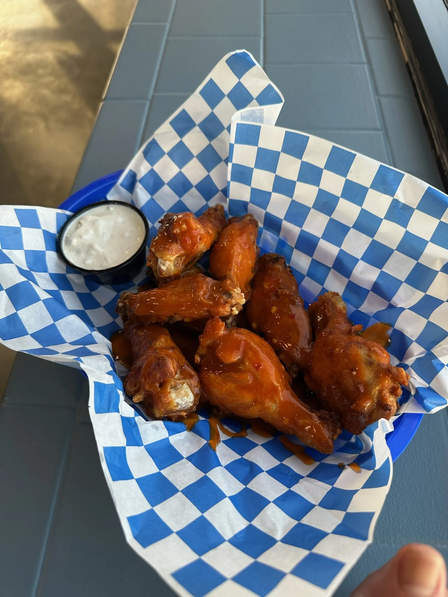 Basket of Cowboy-style chicken wings with a small cup of ranch dipping sauce, placed on blue and white checkered paper at a restaurant, with part of a hand visible in the lower right corner.
