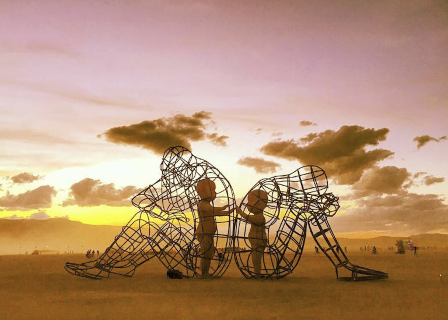 Metal sculpture of a large sitting figure with a smaller figure inside its lap, set against a sunset sky with clouds and distant people.