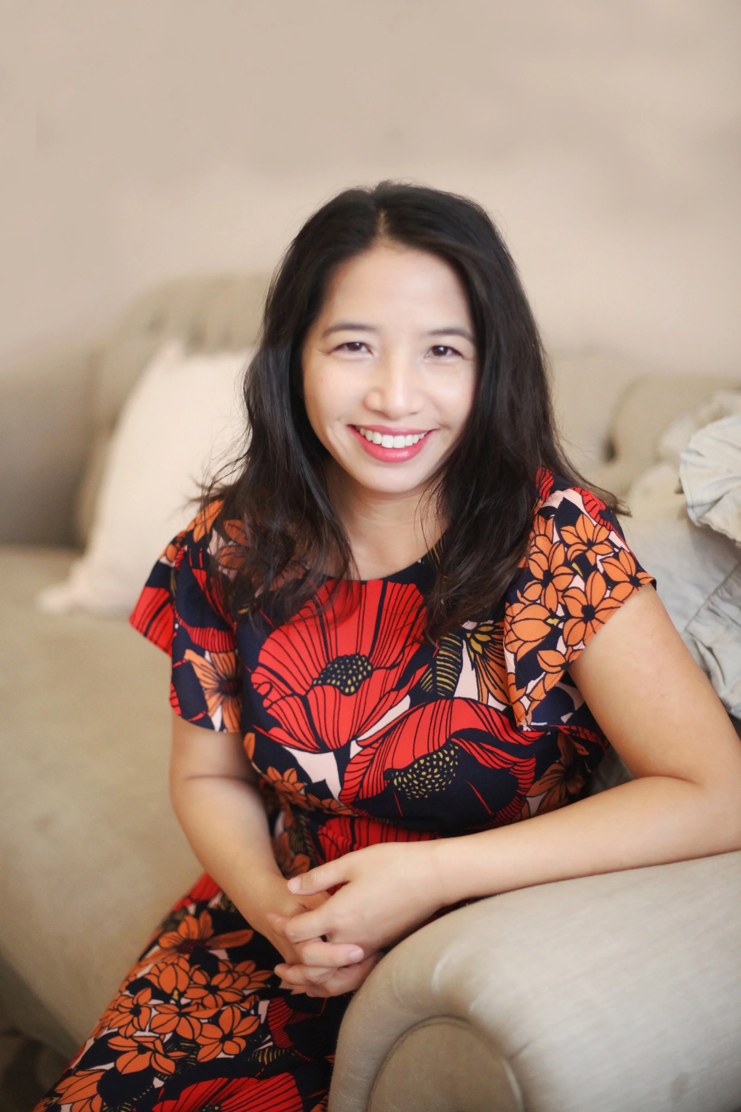 An Asian woman with long dark hair smiling, sitting on a beige sofa in a living room.
