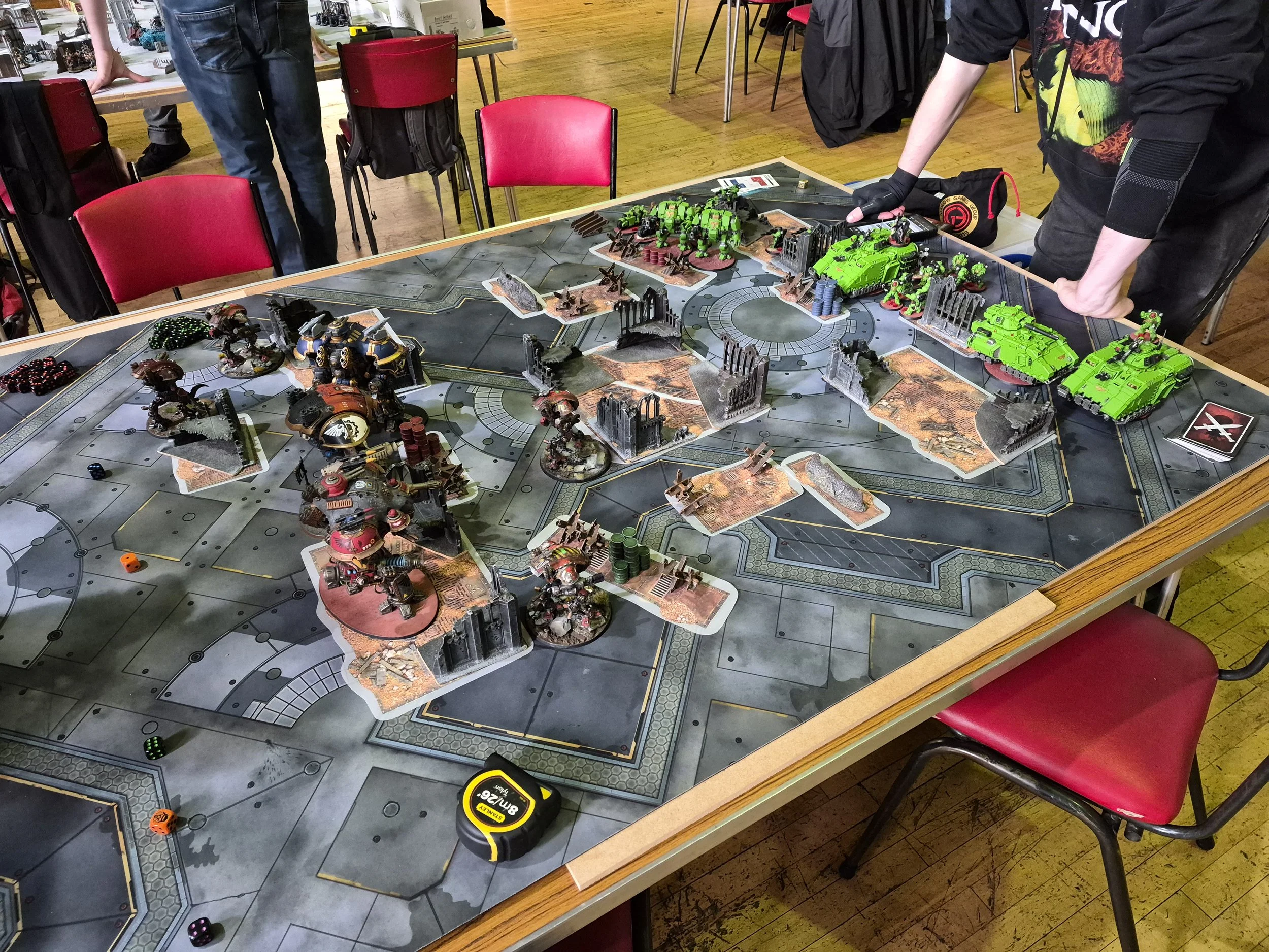A tabletop gaming scene with miniature models resembling futuristic tanks and mechs, arranged on a game board with terrain pieces. Several dice and a tape measure are also visible, and human hands are interacting with the game pieces.
