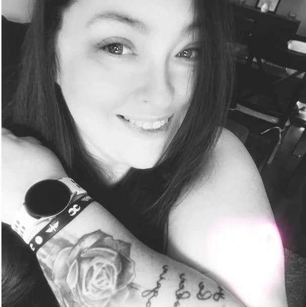 A woman smiling, showing her tattooed arm and wristwatch, with a background of a dining area.