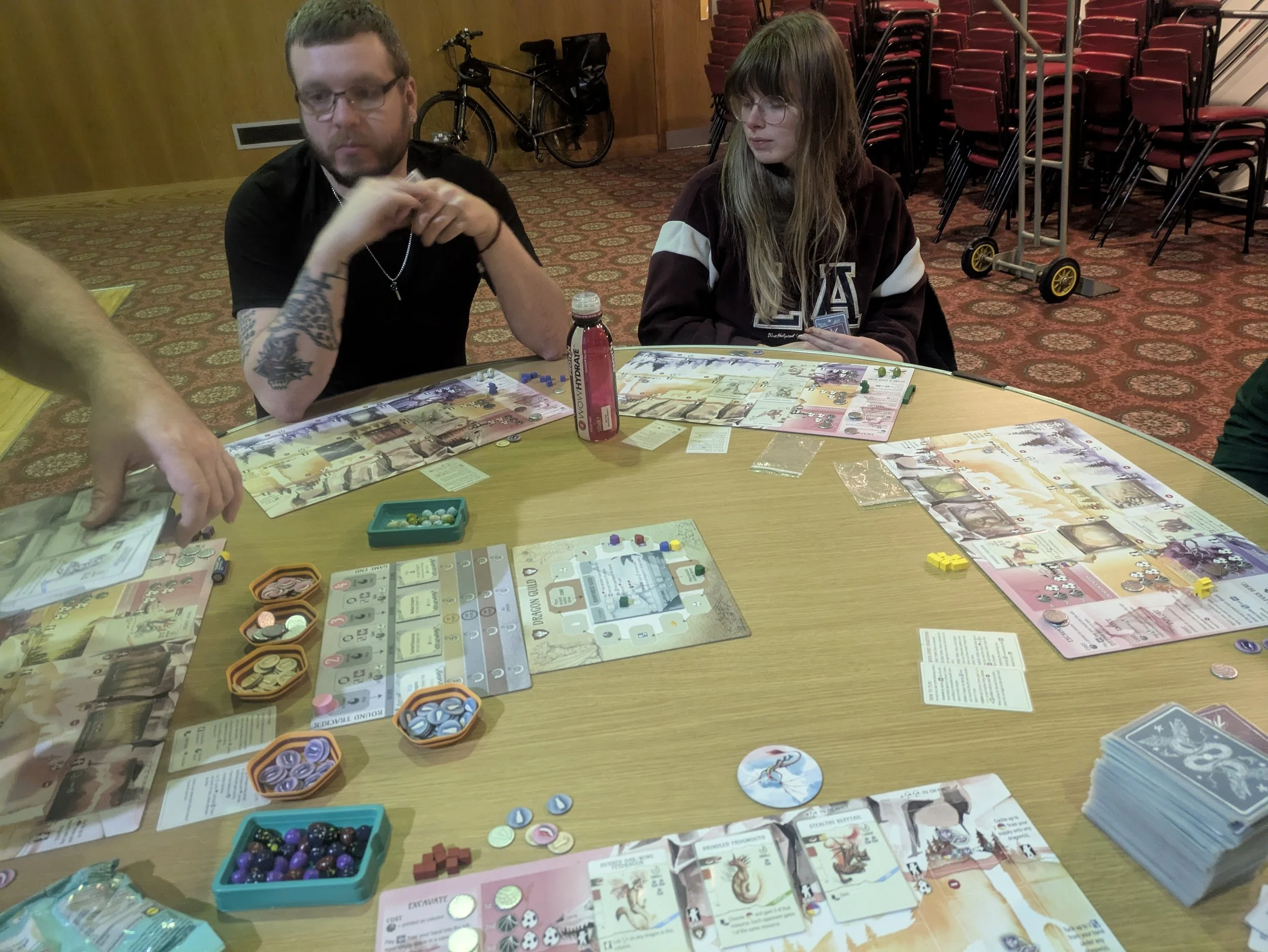 People playing a tabletop board game at a round table in a room with red chairs and stacked chairs in the background.