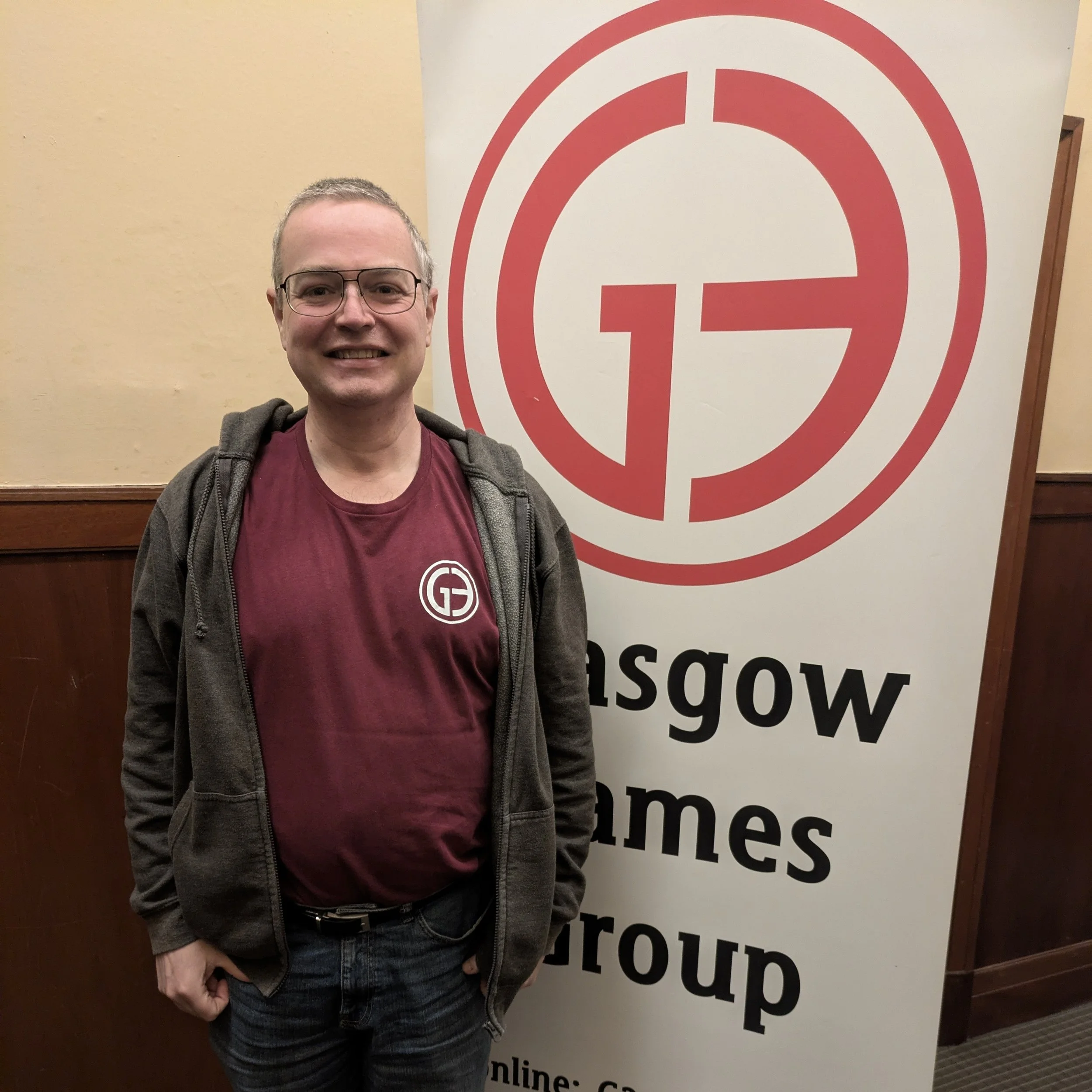 A man wearing glasses, a maroon t-shirt with a white logo, and a gray jacket standing next to a large sign for Glasgow Games Group.