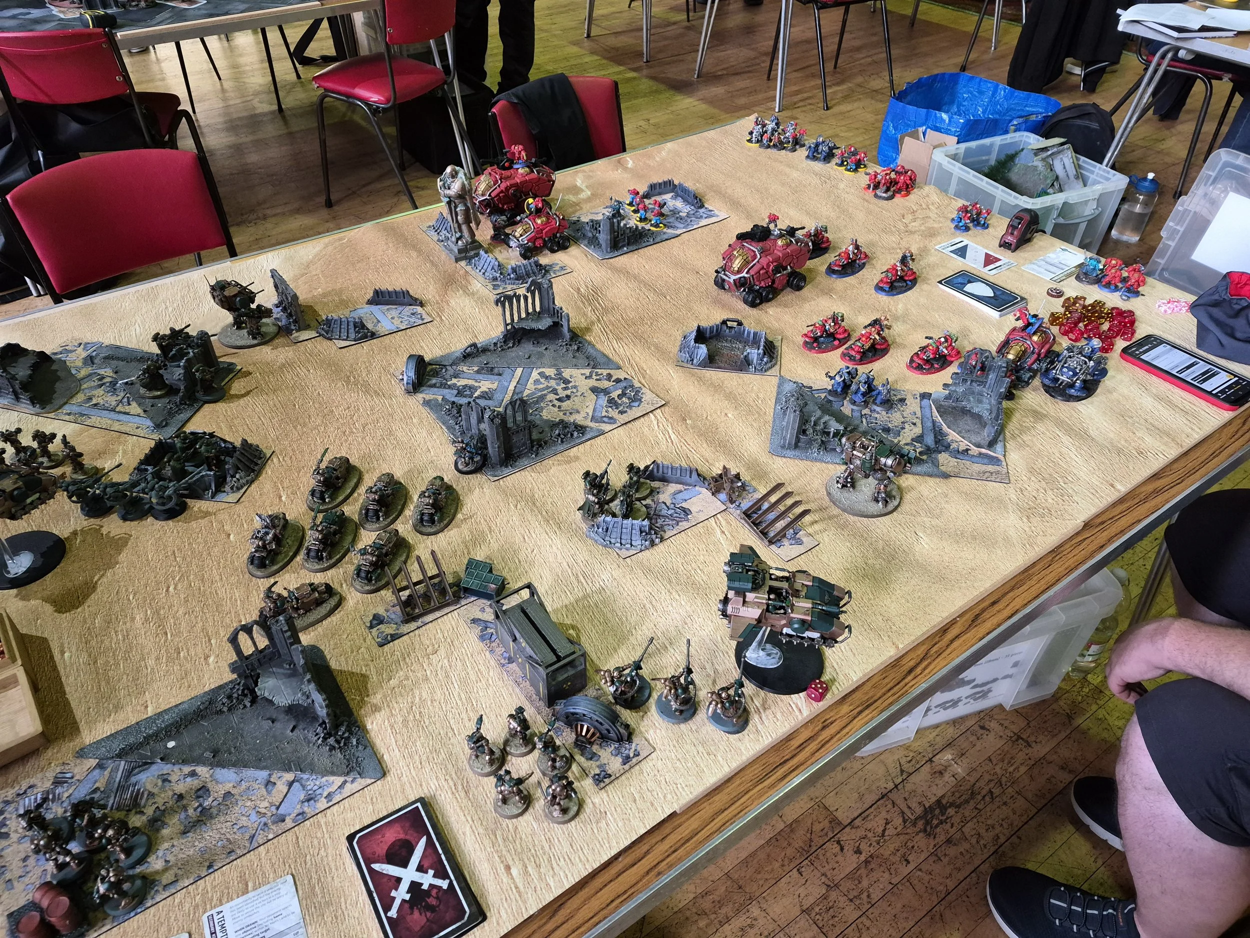 Table with painted miniature models in a tabletop war game setup, surrounded by chairs and various organization containers, with gaming cards, dice, and a smartphone visible.