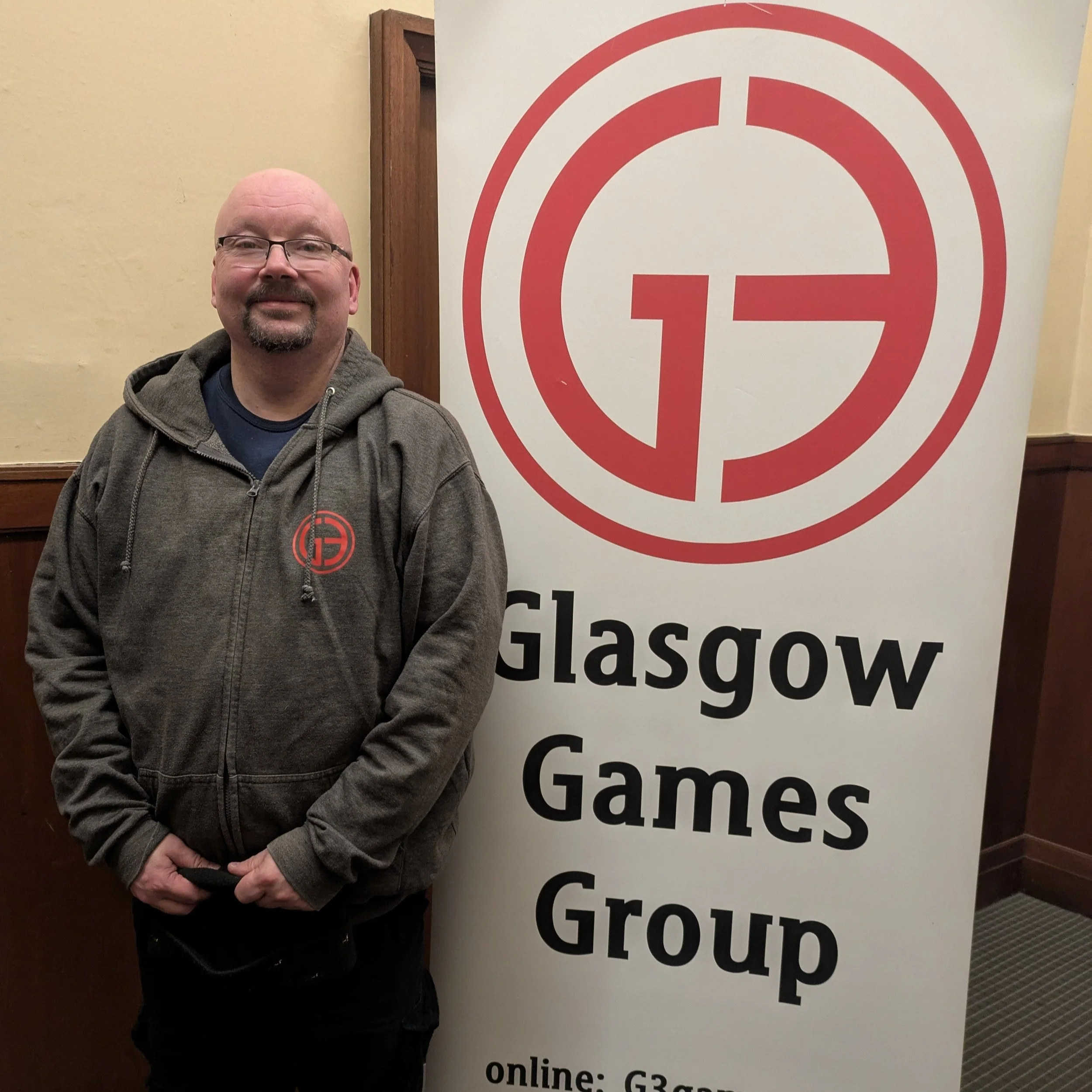 A man wearing a gray hoodie with a Glasgow Games Group logo standing next to a large white banner with the same logo and text.
