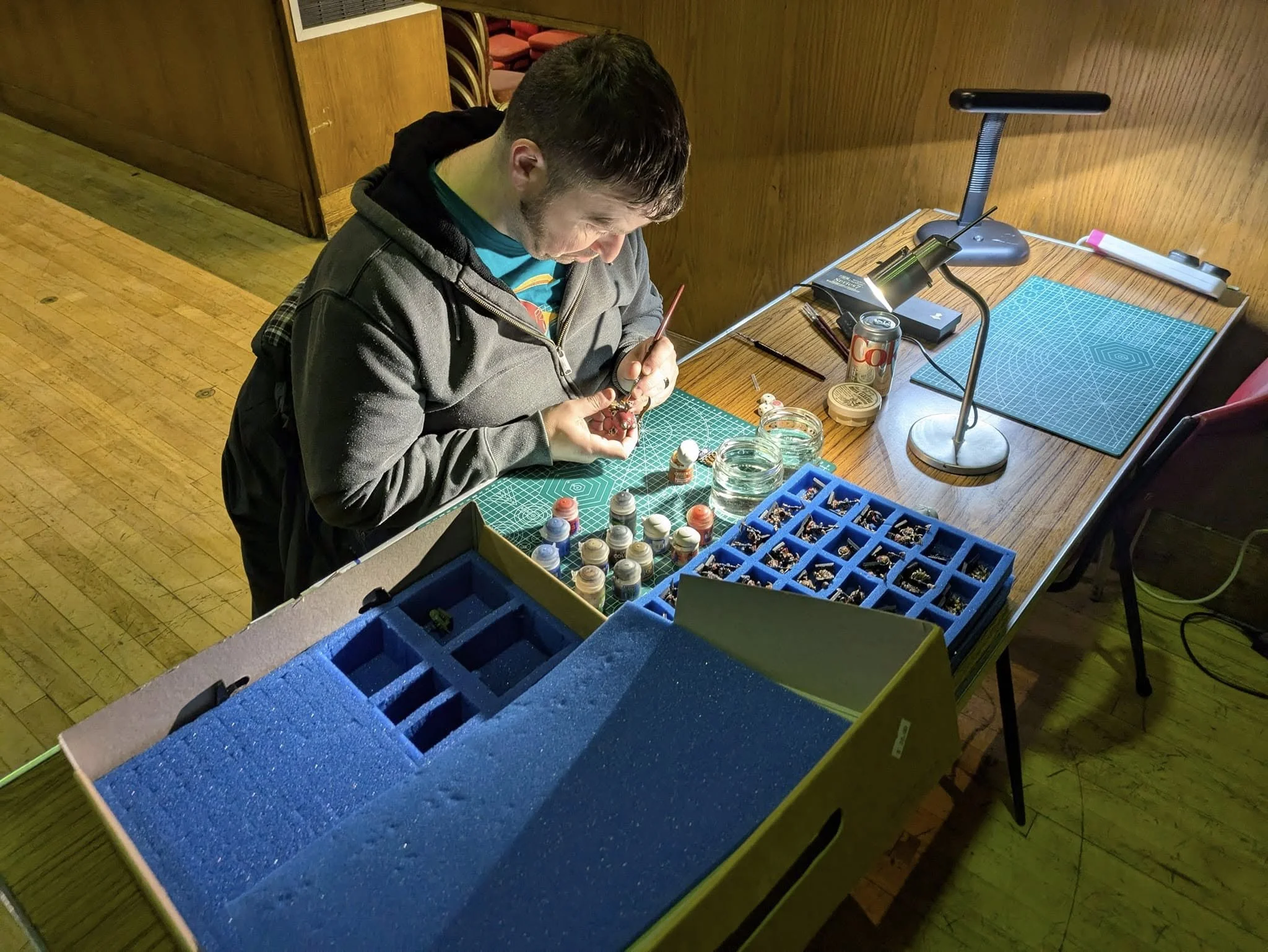 Person organizing miniature models or figures on a crafting table with paints and tools, illuminated by a desk lamp.
