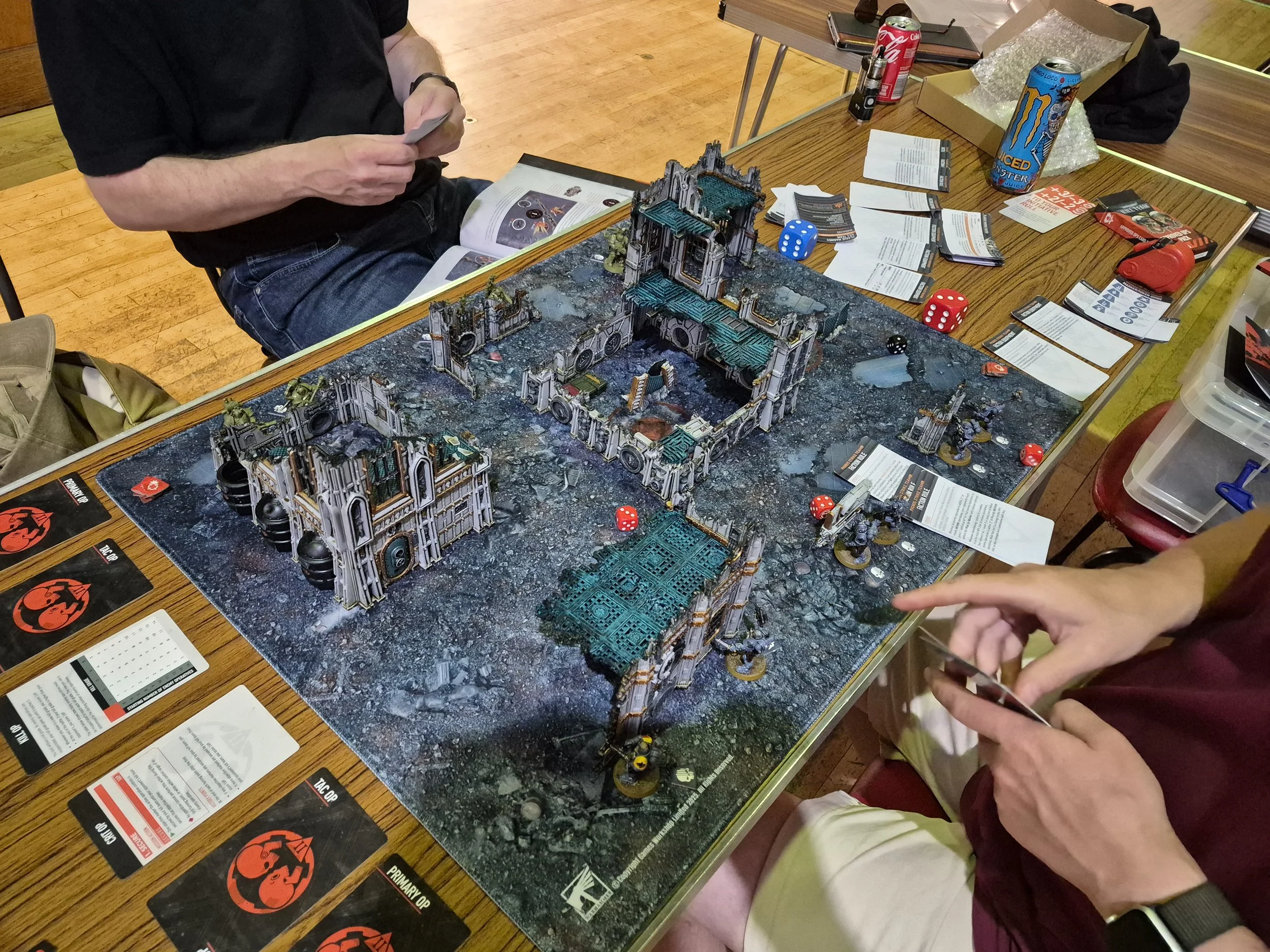 A tabletop role-playing game setup with a detailed terrain and miniatures. Two players are engaged, one holding a card and the other is finger pointing. The table has various cards, dice, beverages, and rulebooks. The terrain includes ruins and structures resembling a ruined city or sci-fi setting.