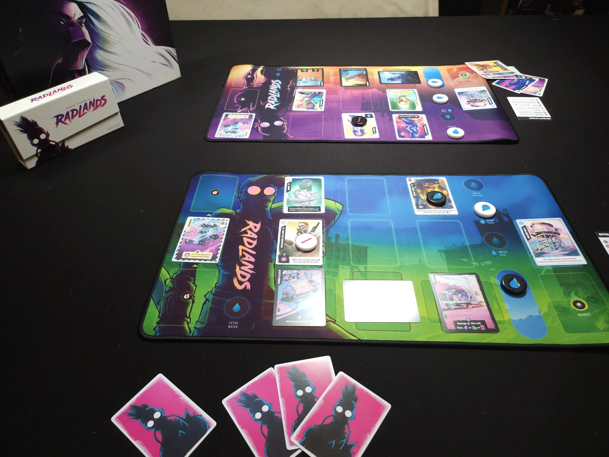 Two board game tables set up with colorful game boards, playing cards, tokens, and game pieces, with a box and artwork from the game Radlands visible in the background.