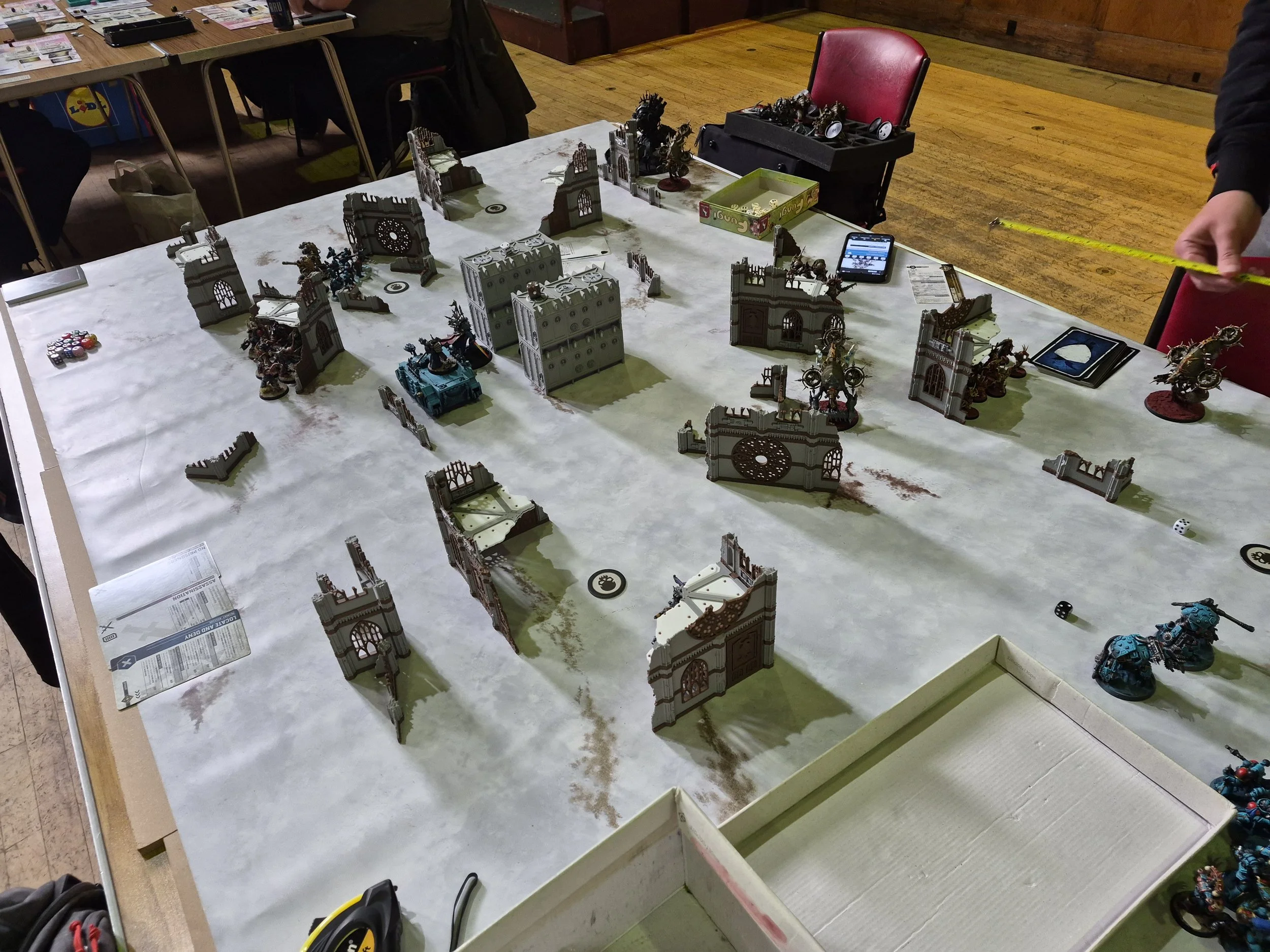 Tabletop miniature war game with detailed miniature buildings, vehicles, and figures, set on a white textured gaming surface in a room with wooden floors and red chairs.