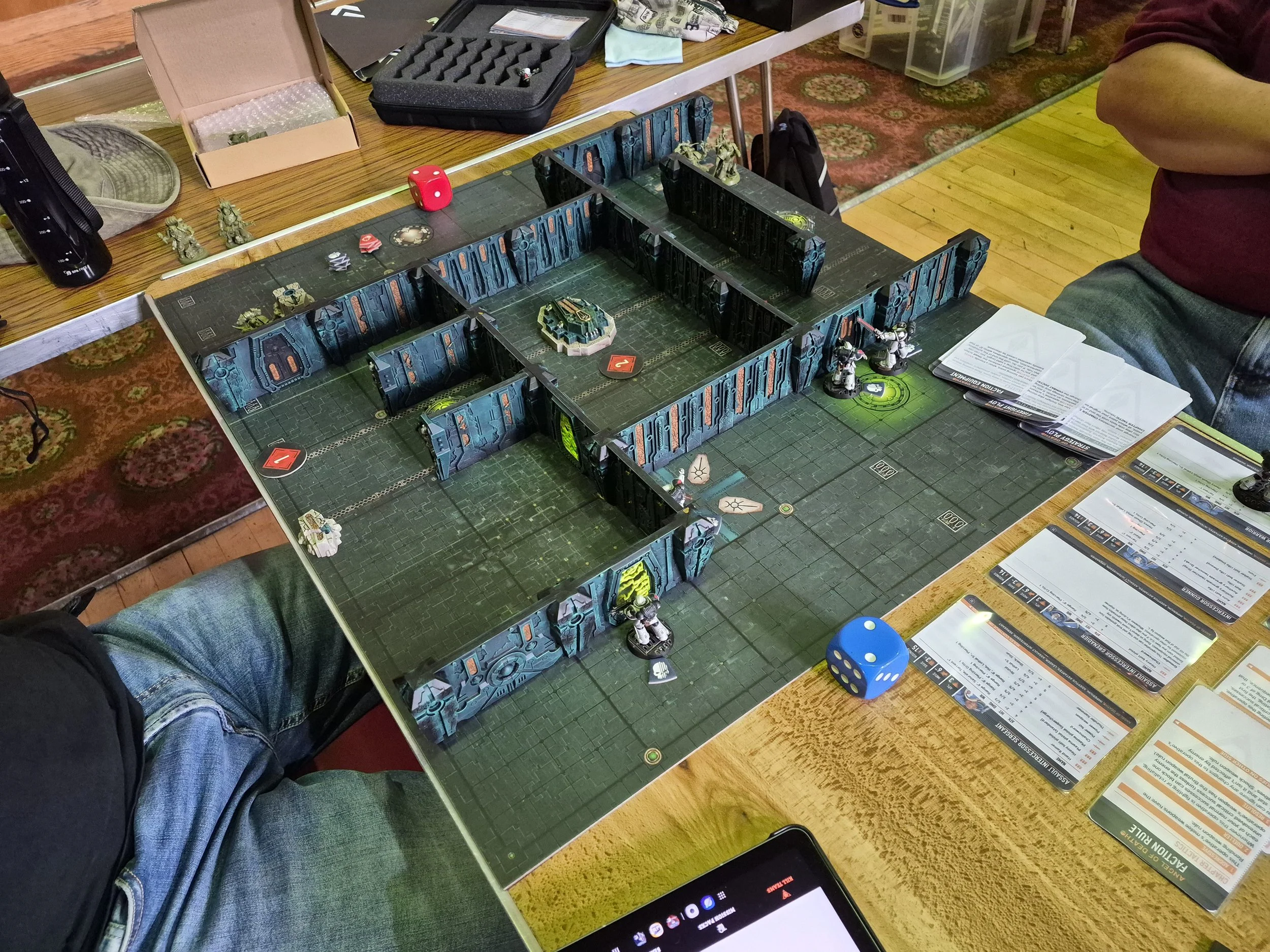 Tabletop role-playing game setup with modular game board, miniatures, dice, and rule books. Player's arm and leg visible, game accessories on surrounding tables and carpeted floor.