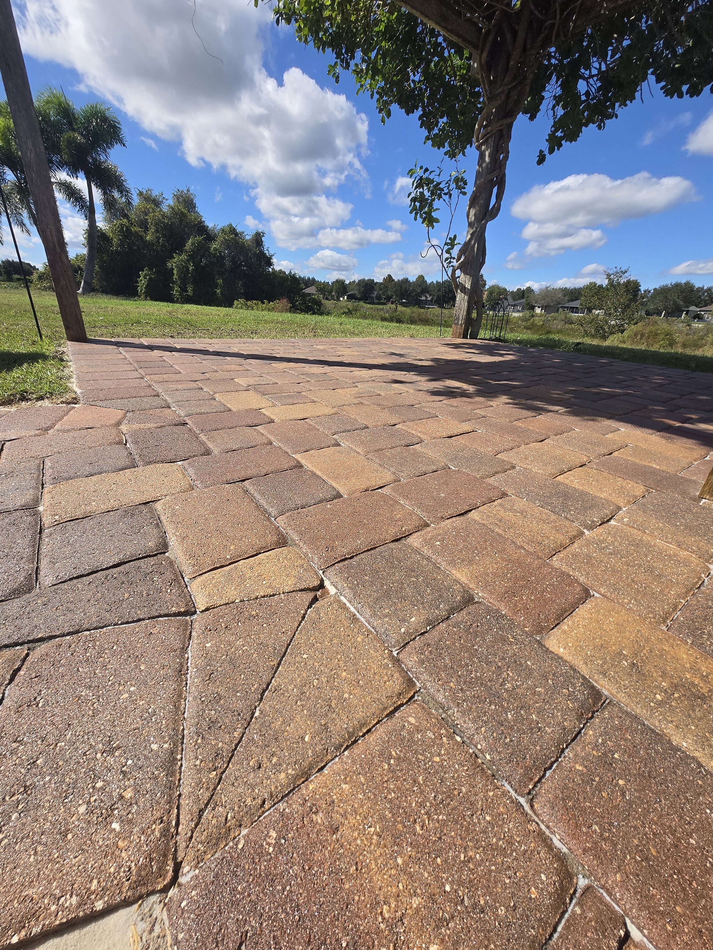 Close-up of a brick patio under a shaded pergola with green grass, trees, and a cloudy blue sky in the background.