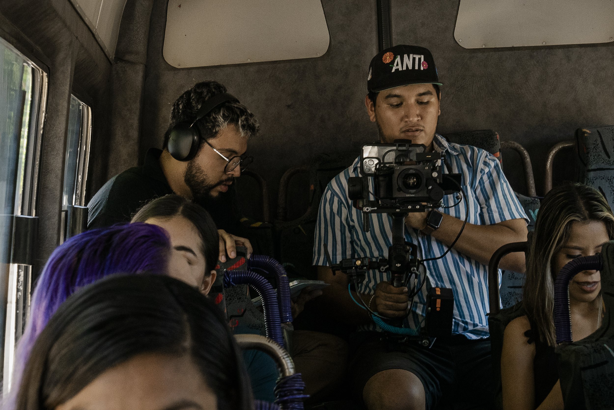 Equipo de filmación en un autobús, con un camarógrafo usando un estabilizador de cámara y varias personas sentadas con equipo de audio y accesorios relacionados con la filmación.