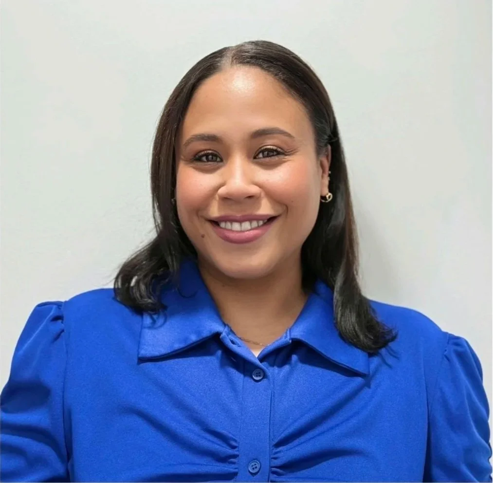 A woman with medium-length dark hair, smiling, wearing a royal blue button-up shirt, standing against a plain white wall.