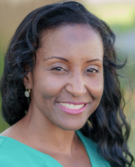 Smiling woman with dark curly hair, wearing a green top and earrings, outdoors.